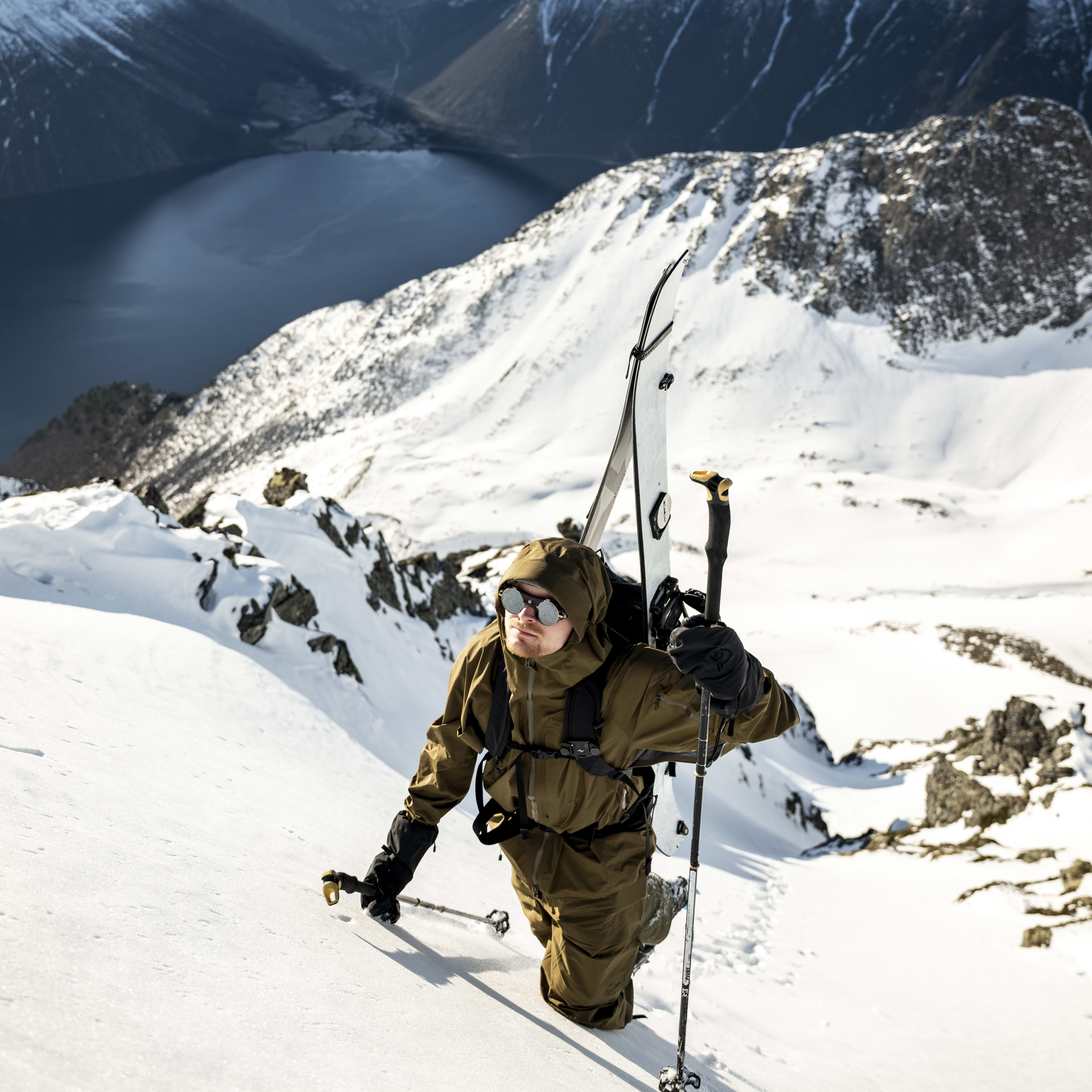 A man in Norrøna outdoor gear climbs a snowy mountain with skis on his back. Mountain range and lake in background.