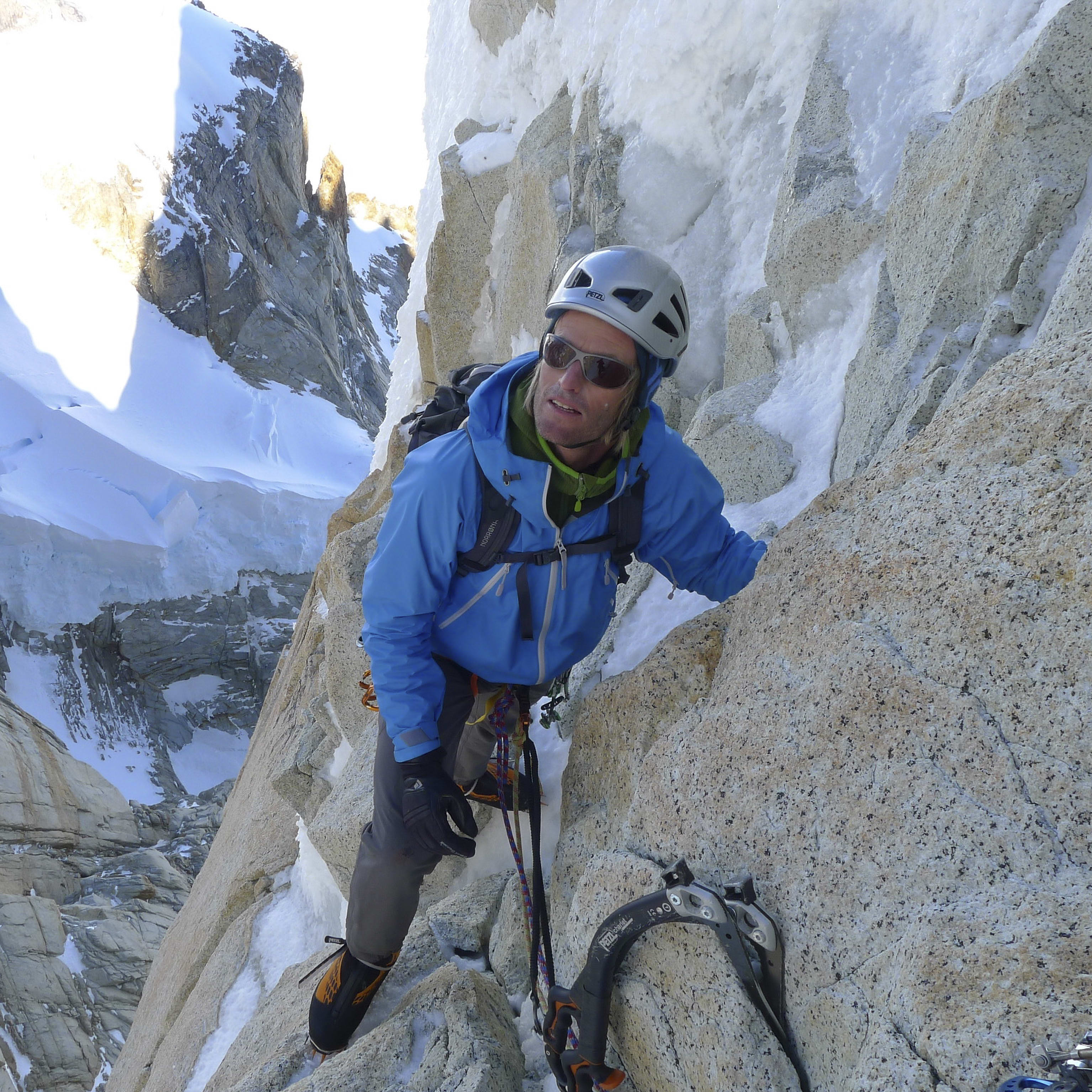 A rock climber wearing a helmet and a blue jacket is climbing a snowy mountain face. The climber is using a technical ice axe and has a rope attached to their harness. The background shows a large snowy mountain face.