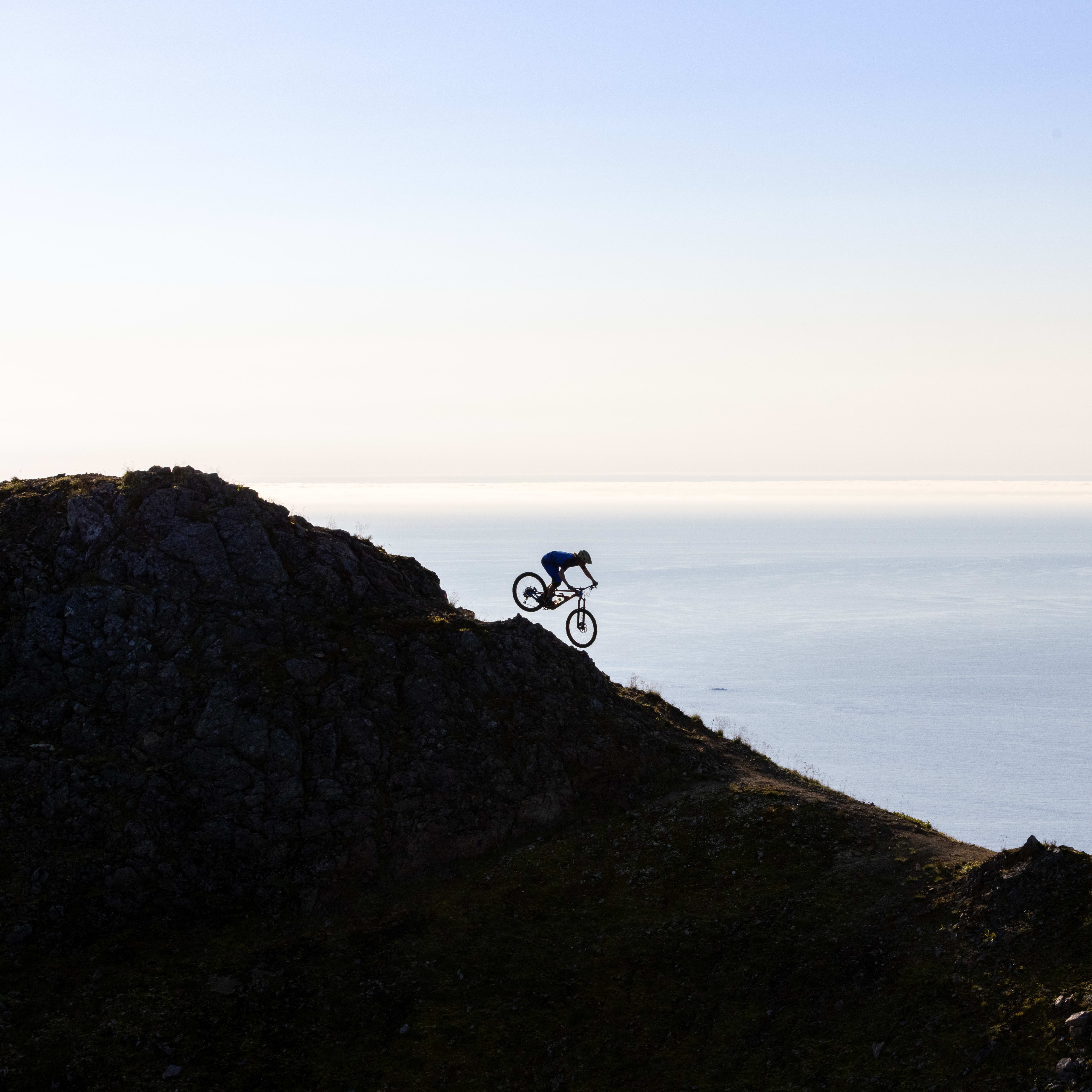 Silhouette of a person riding a Norrøna mountain bike down a rocky cliff overlooking the ocean. 
