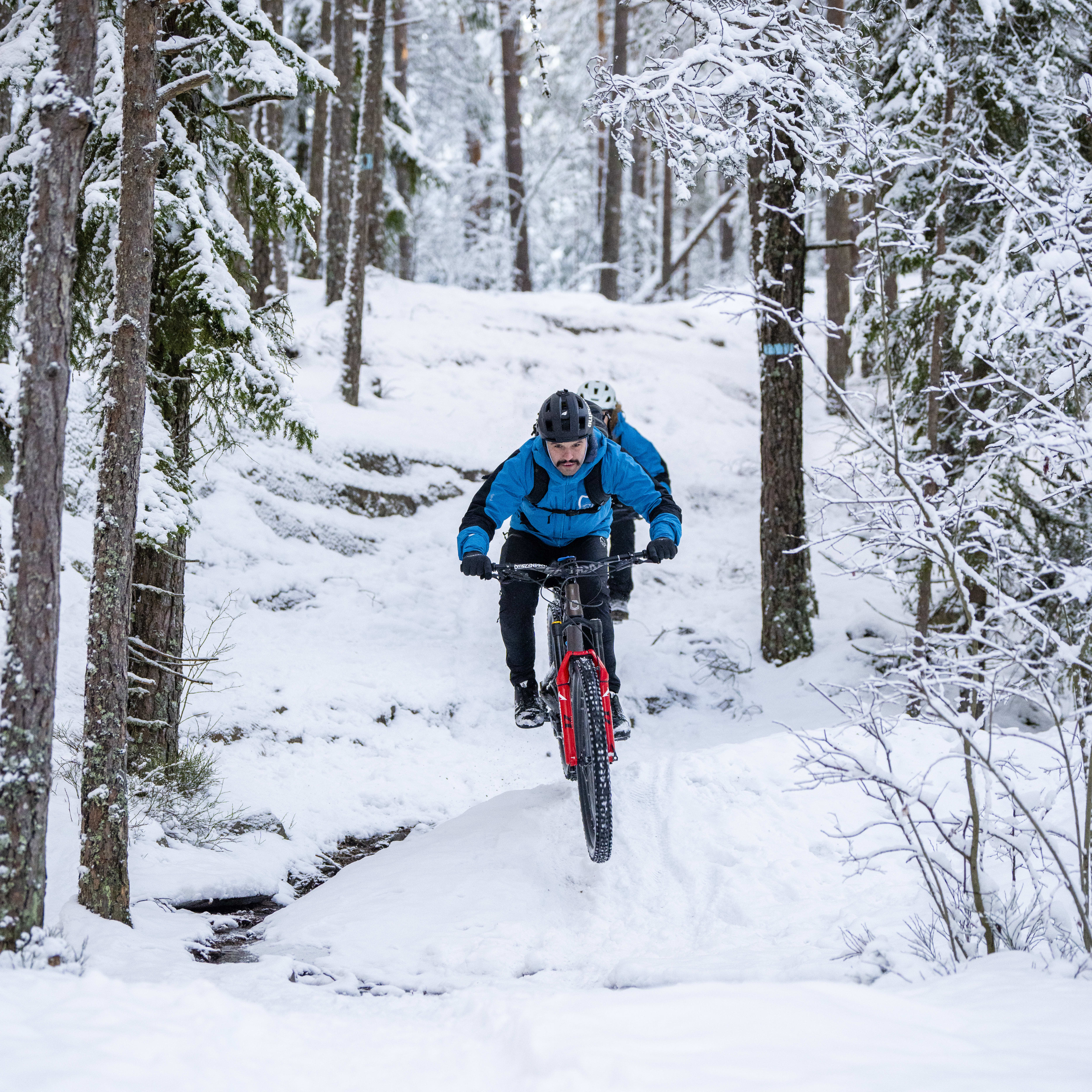 A person wearing a Norrøna jacket rides a mountain bike up a snowy trail in a forest.  
