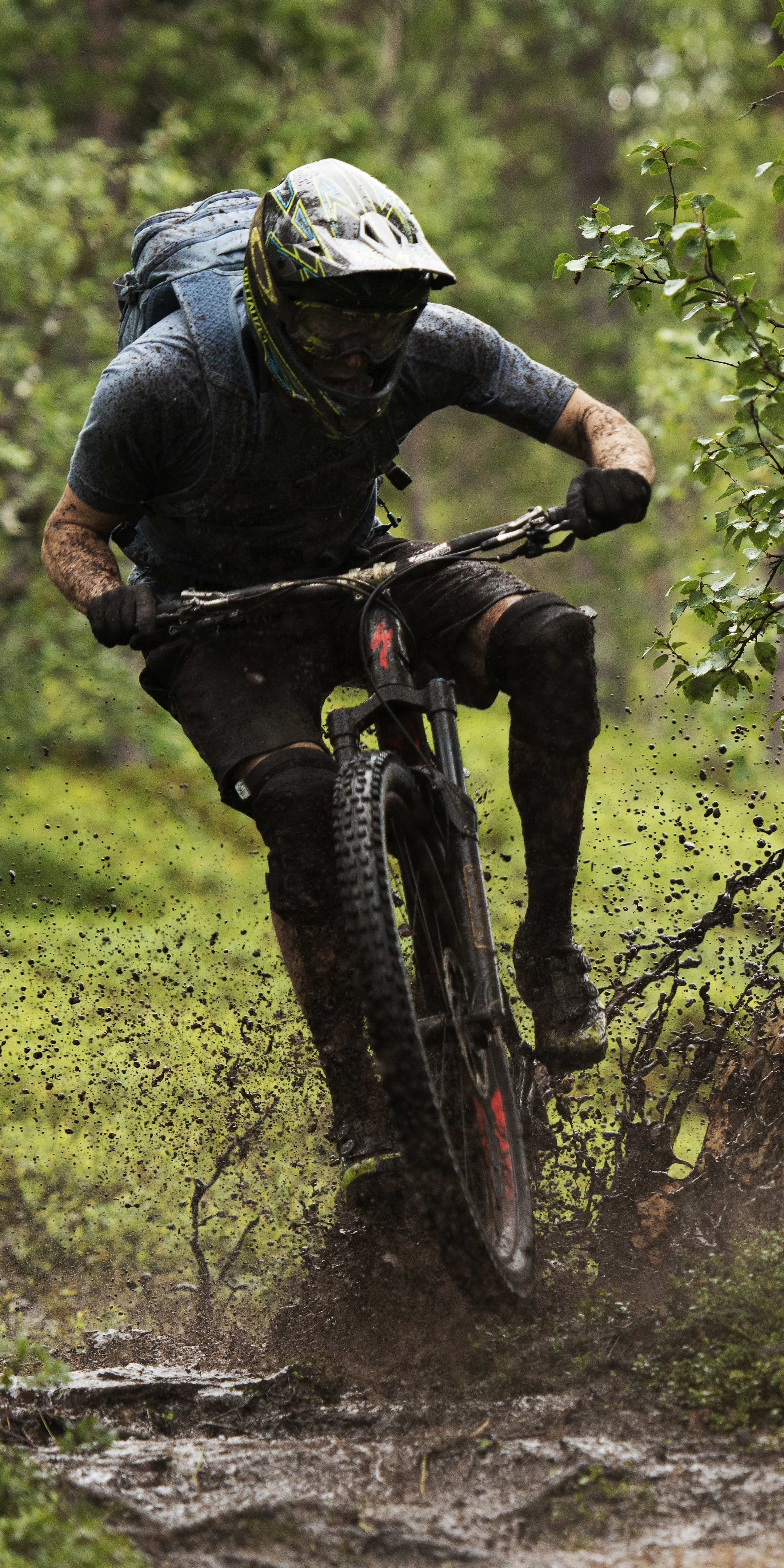 A person in a Norrøna cycling outfit rides a mountain bike through a muddy forest path, kicking up a spray of dirt.