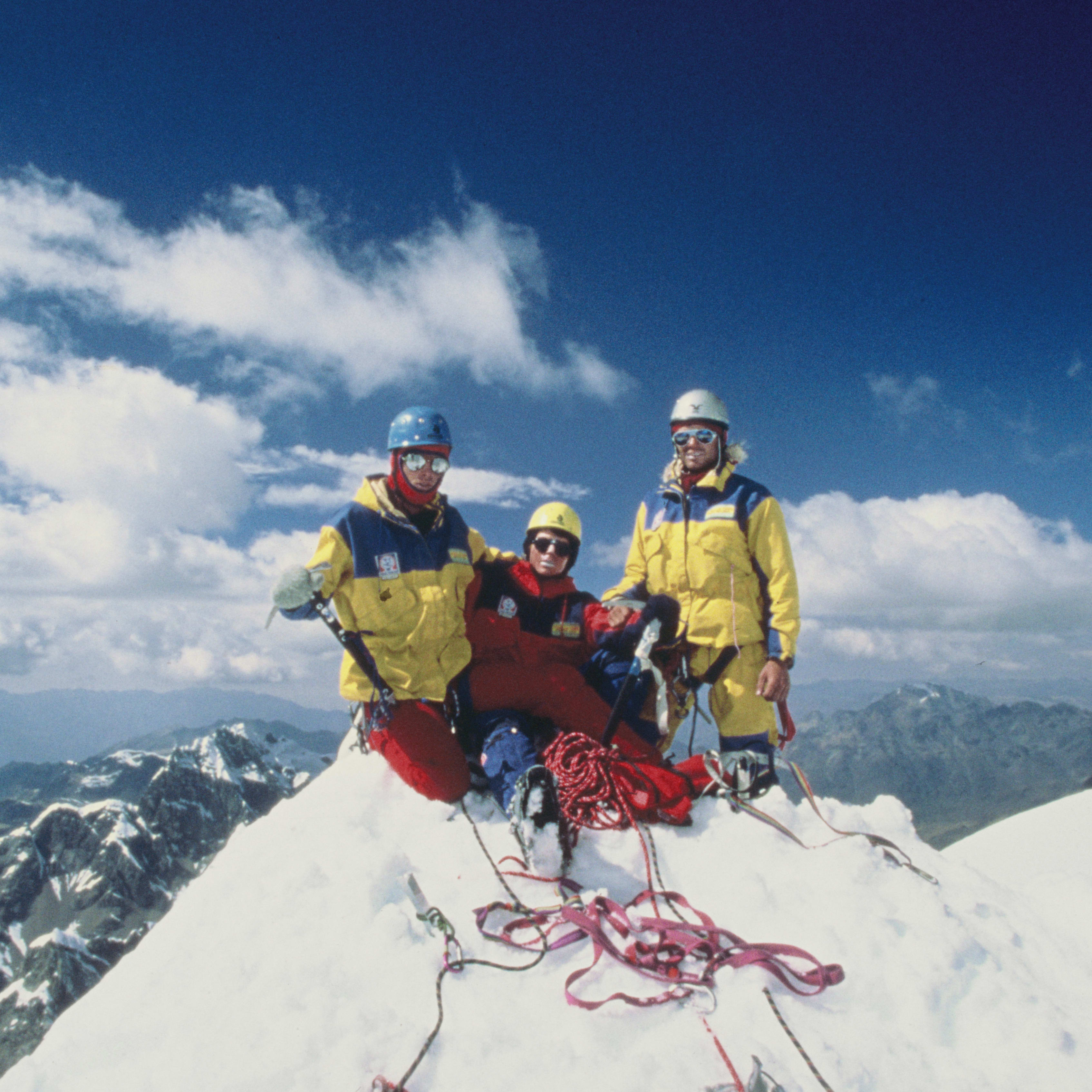 Three mountain climbers stand on a snow-covered peak, smiling at the camera. A blue sky with white clouds is behind them. There are mountains in the distance. The climbers are wearing warm clothing and helmets. One climber is sitting down. There is a red rope and other climbing equipment on the snow around them.