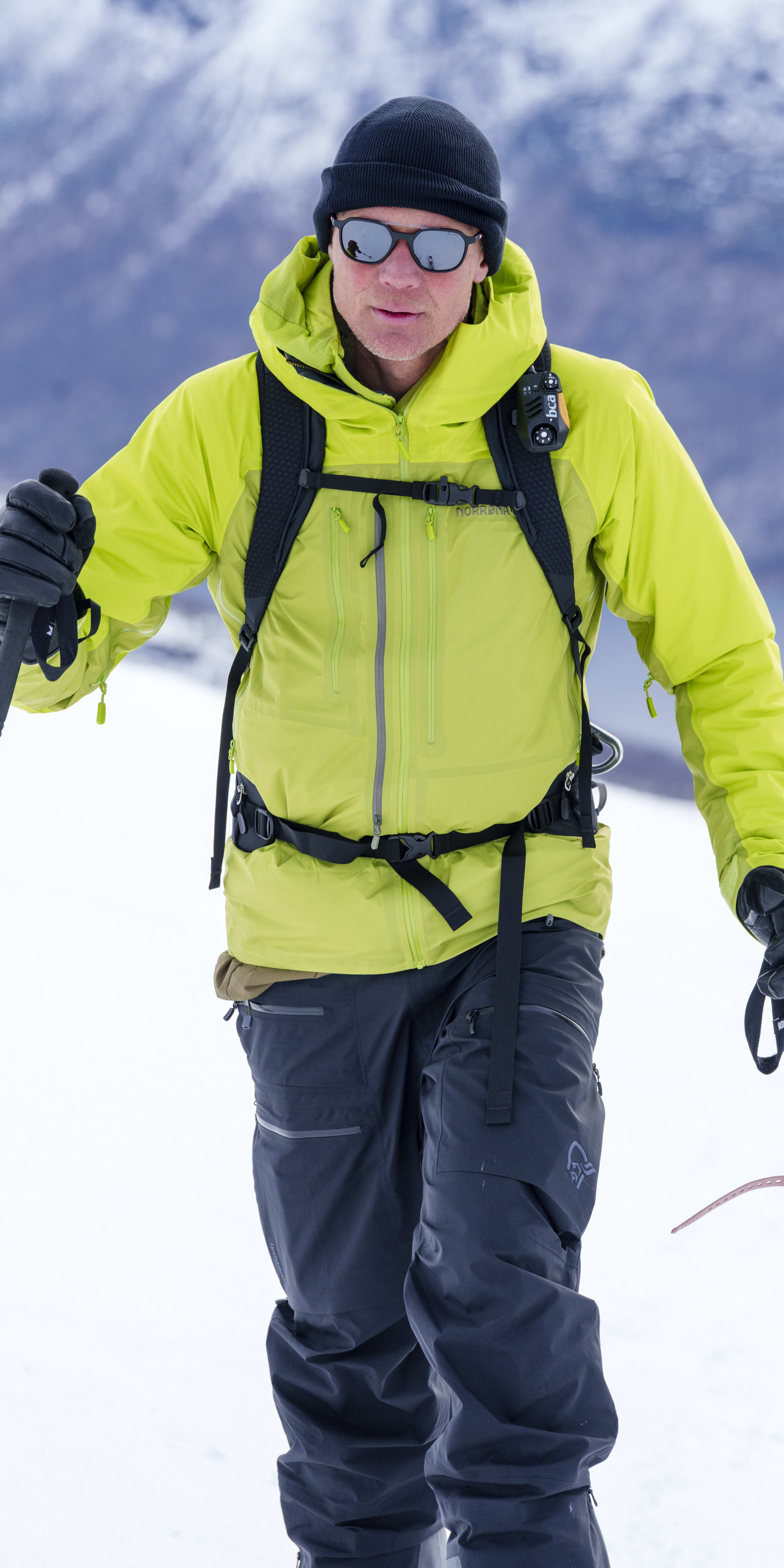 A skier in the mountains. He wears a black beanie, sunglasses, a bright yellow Norrøna jacket, and gray pants. He holds ski poles.