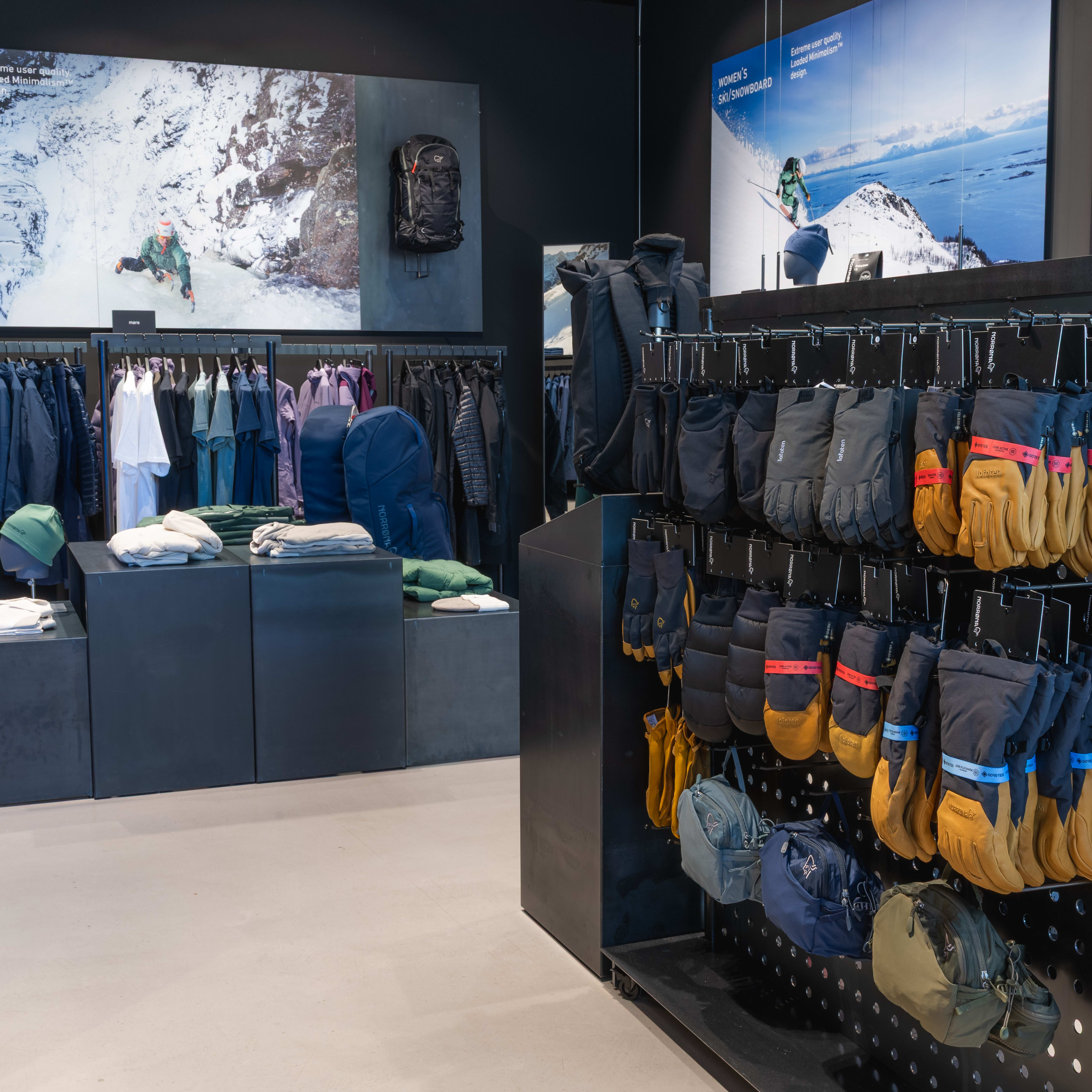 A retail space displays Norrøna clothing and accessories. Racks hold garments and gloves; display cases feature folded items and backpacks. Wall graphics show winter landscapes.