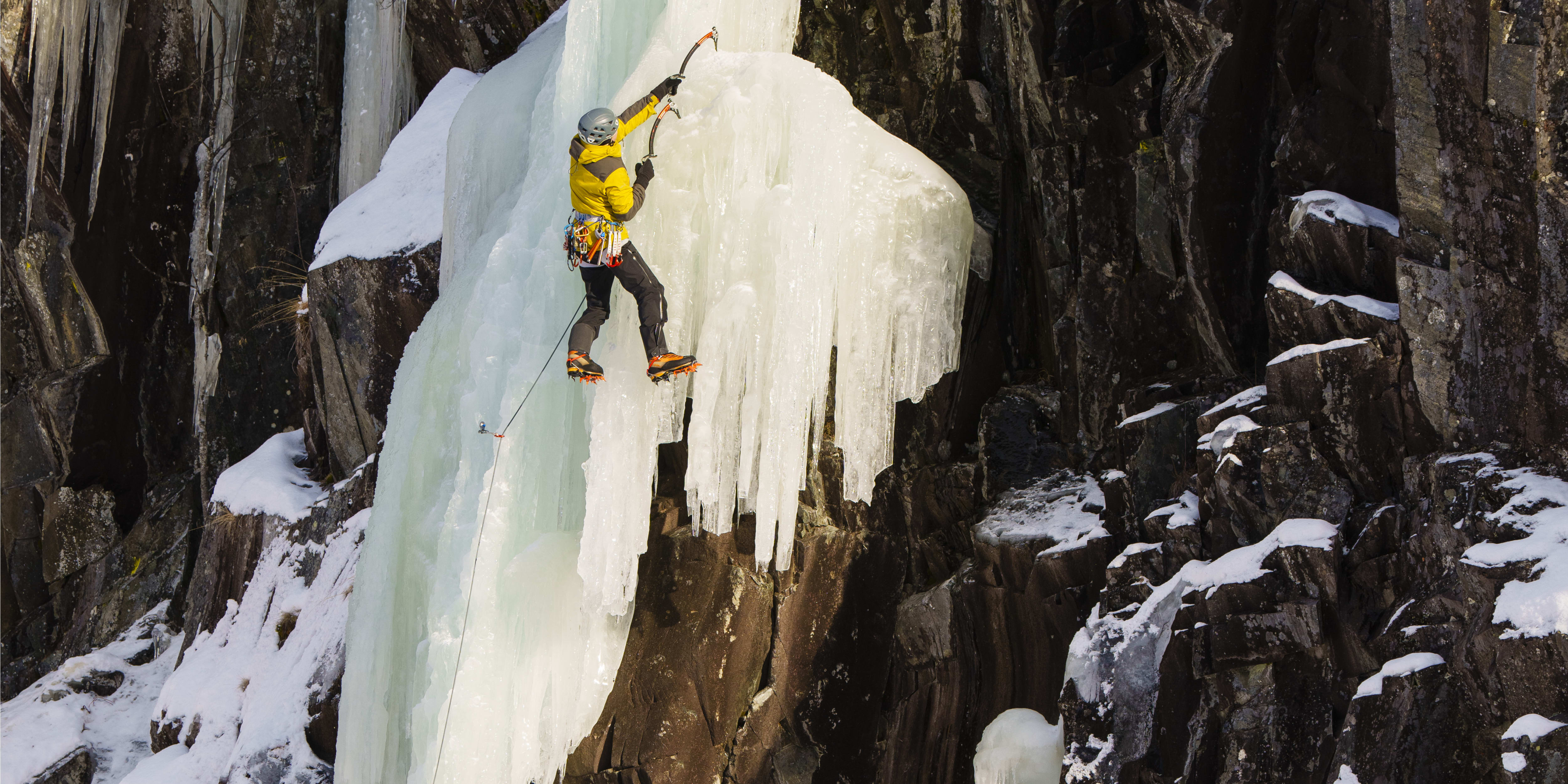 A person ice climbing a frozen waterfall. The climber wears a yellow Norrøna jacket and a helmet.