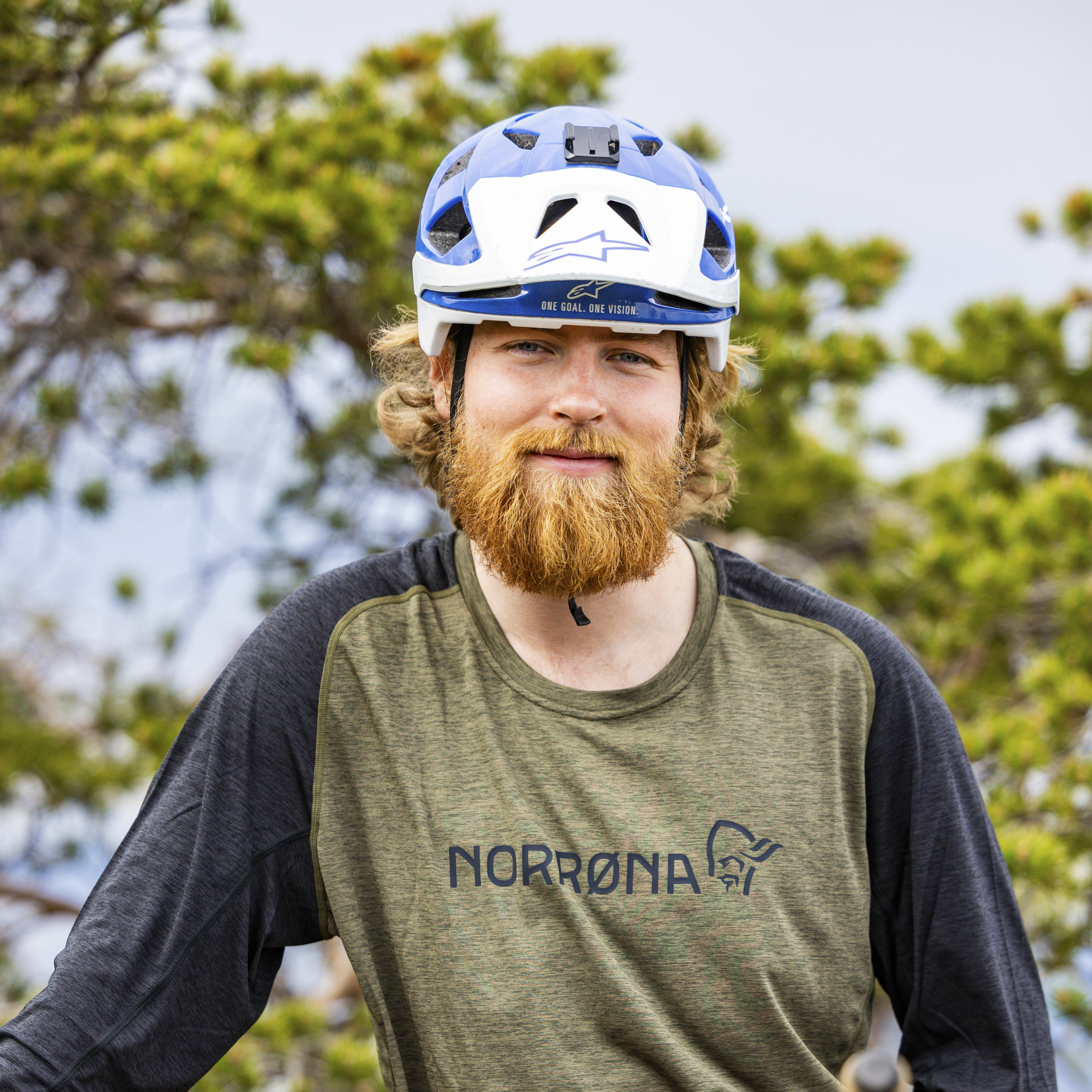A man with a beard wearing a Norrøna shirt and a helmet stands next to a mountain bike.