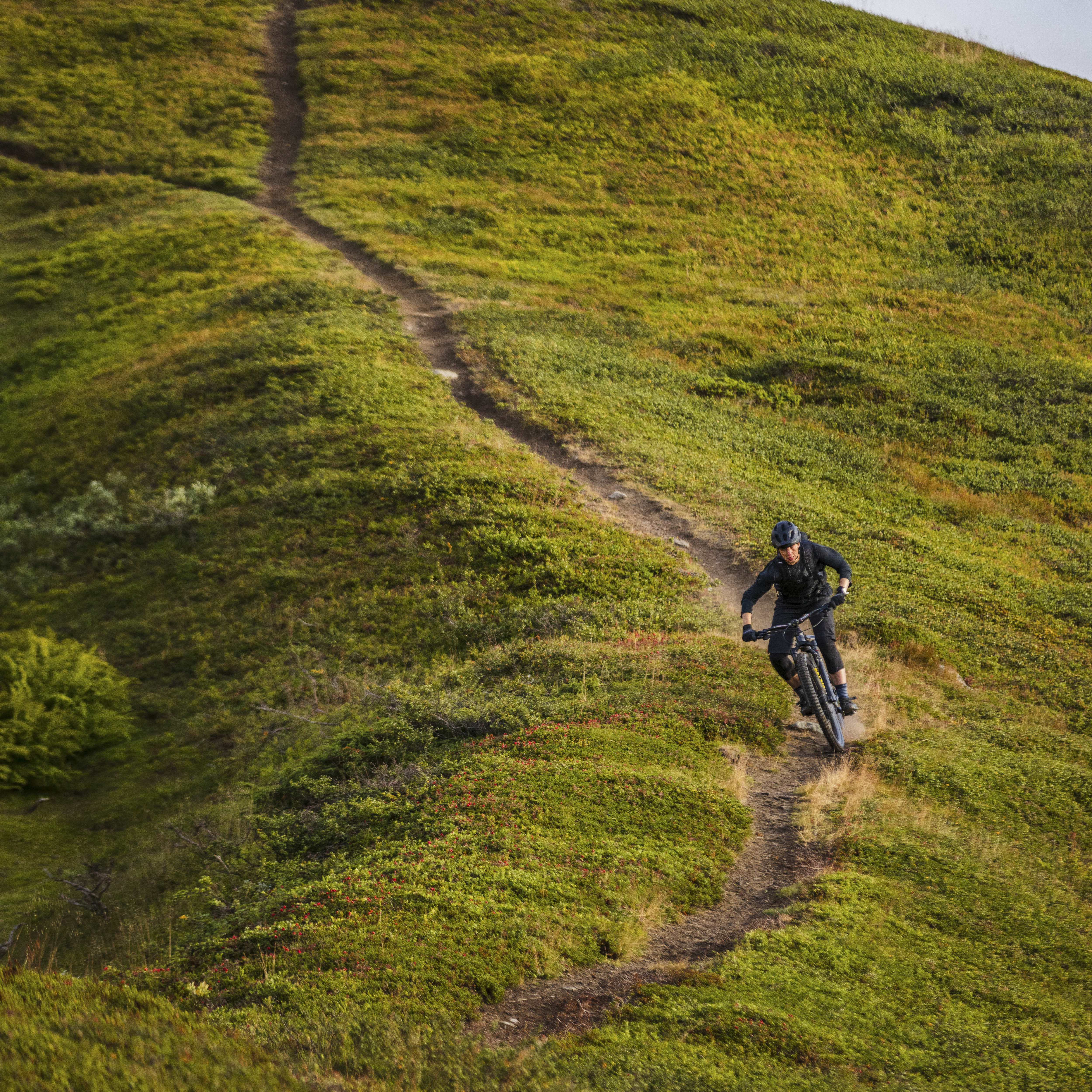 A mountain biker in Norrøna clothing descends a grassy hillside trail. The path winds up to a rocky summit against a bright sky.