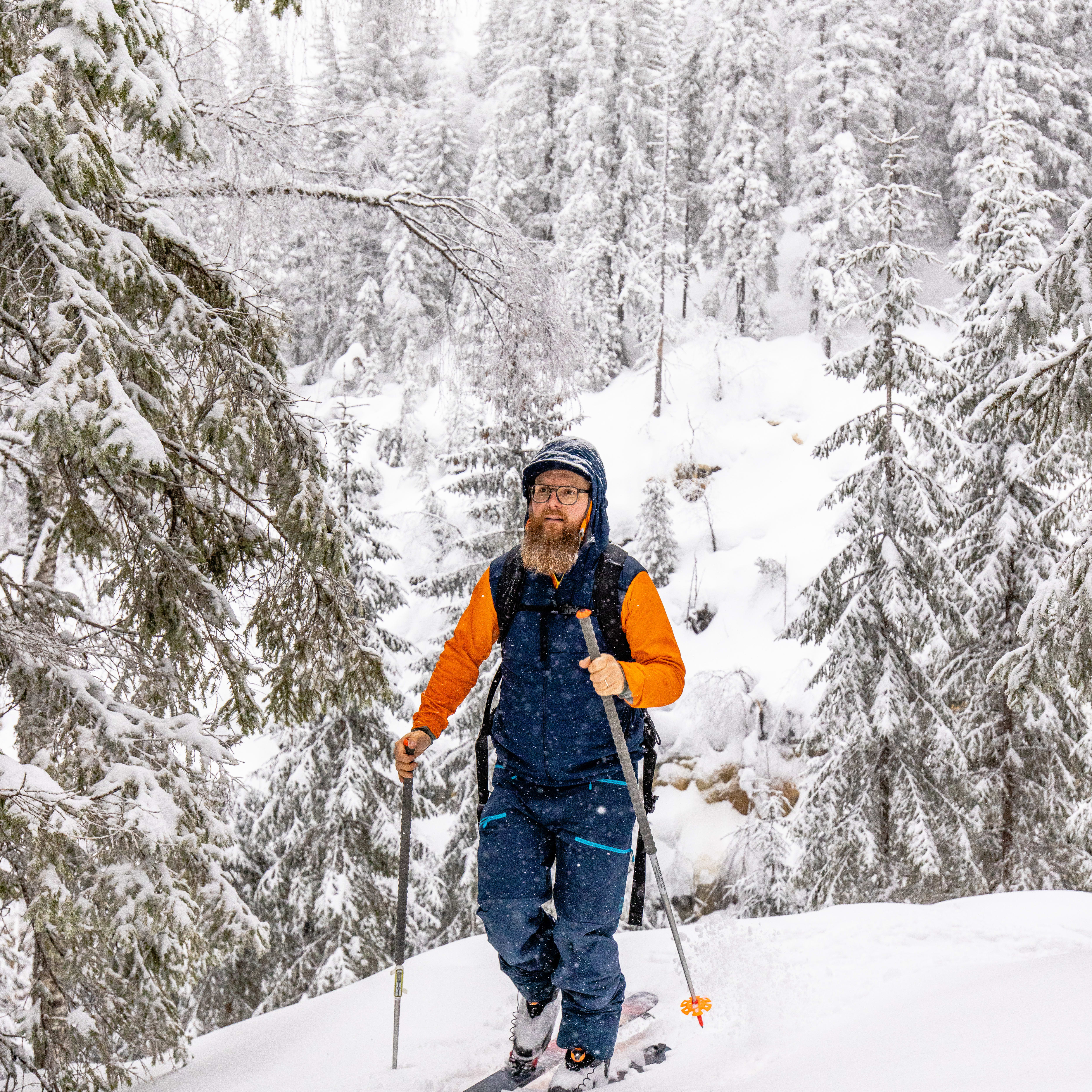 A man in Norrøna clothing backcountry skis through a snowy forest. 
