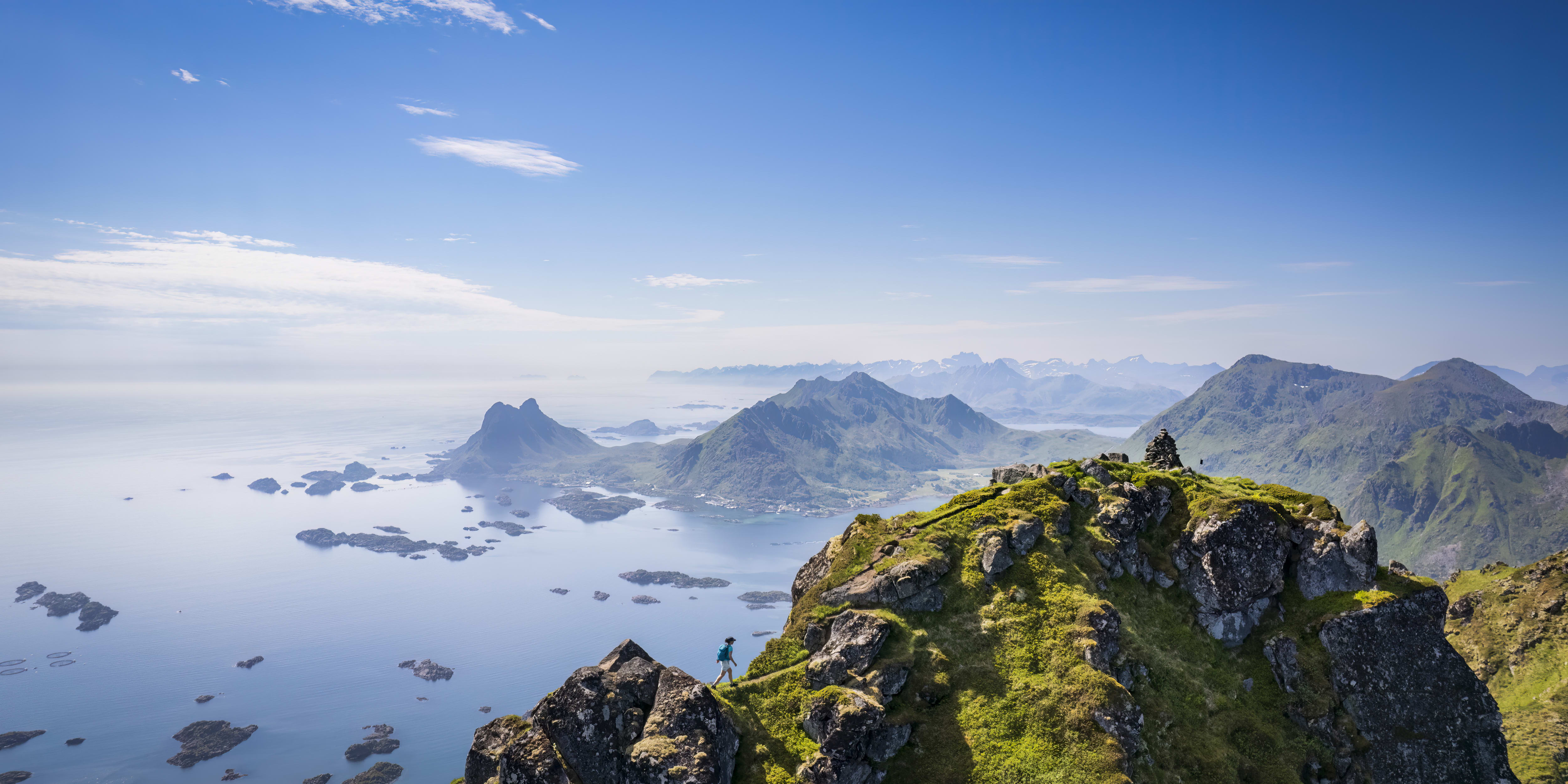 A person stands on a mountaintop overlooking a blue ocean with many rocky islands. The horizon is a hazy blue and the sky is clear and blue.