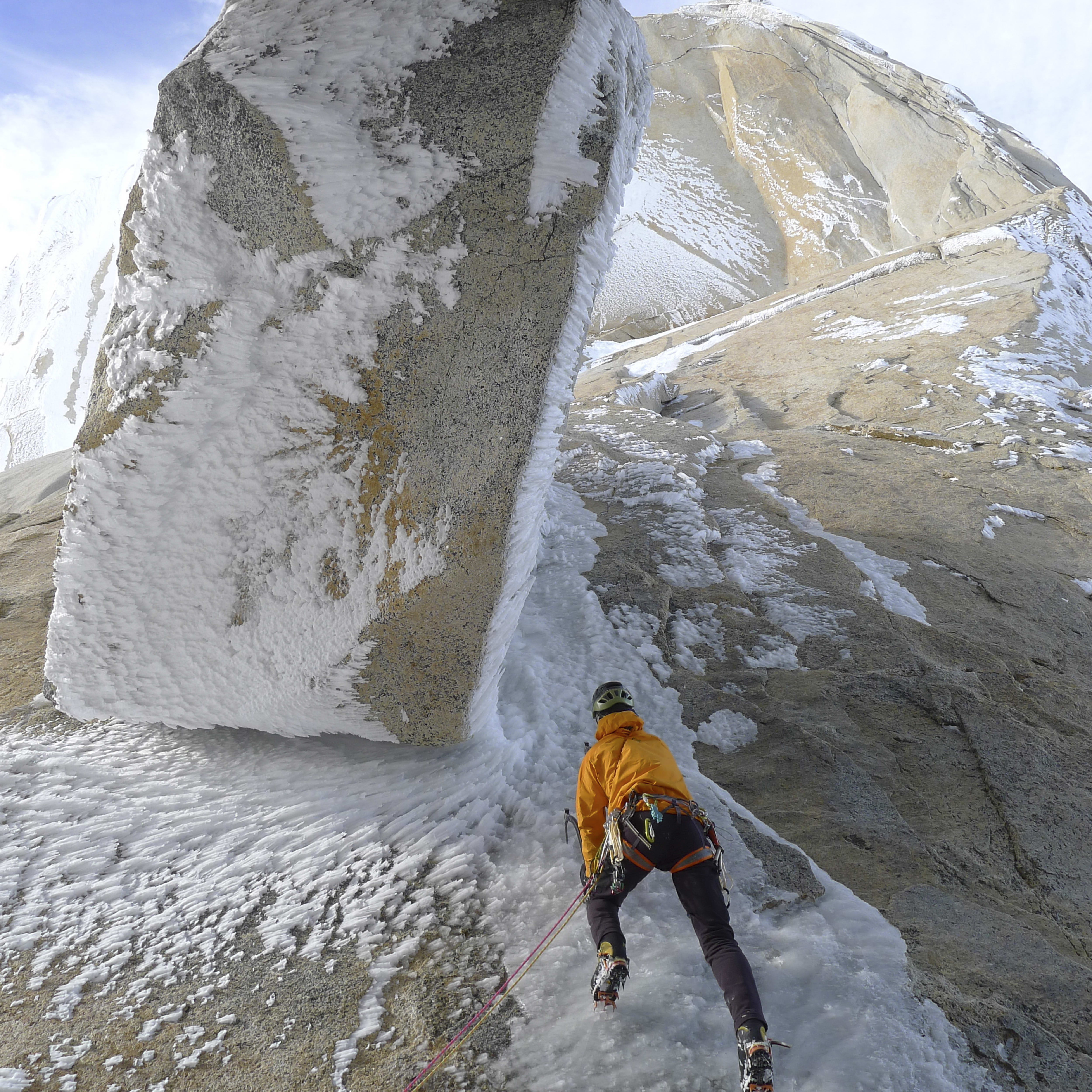 A rock climber in an orange jacket is climbing a snow and ice-covered rock face. The climber is wearing crampons. The blue sky is visible through the icy overhang.