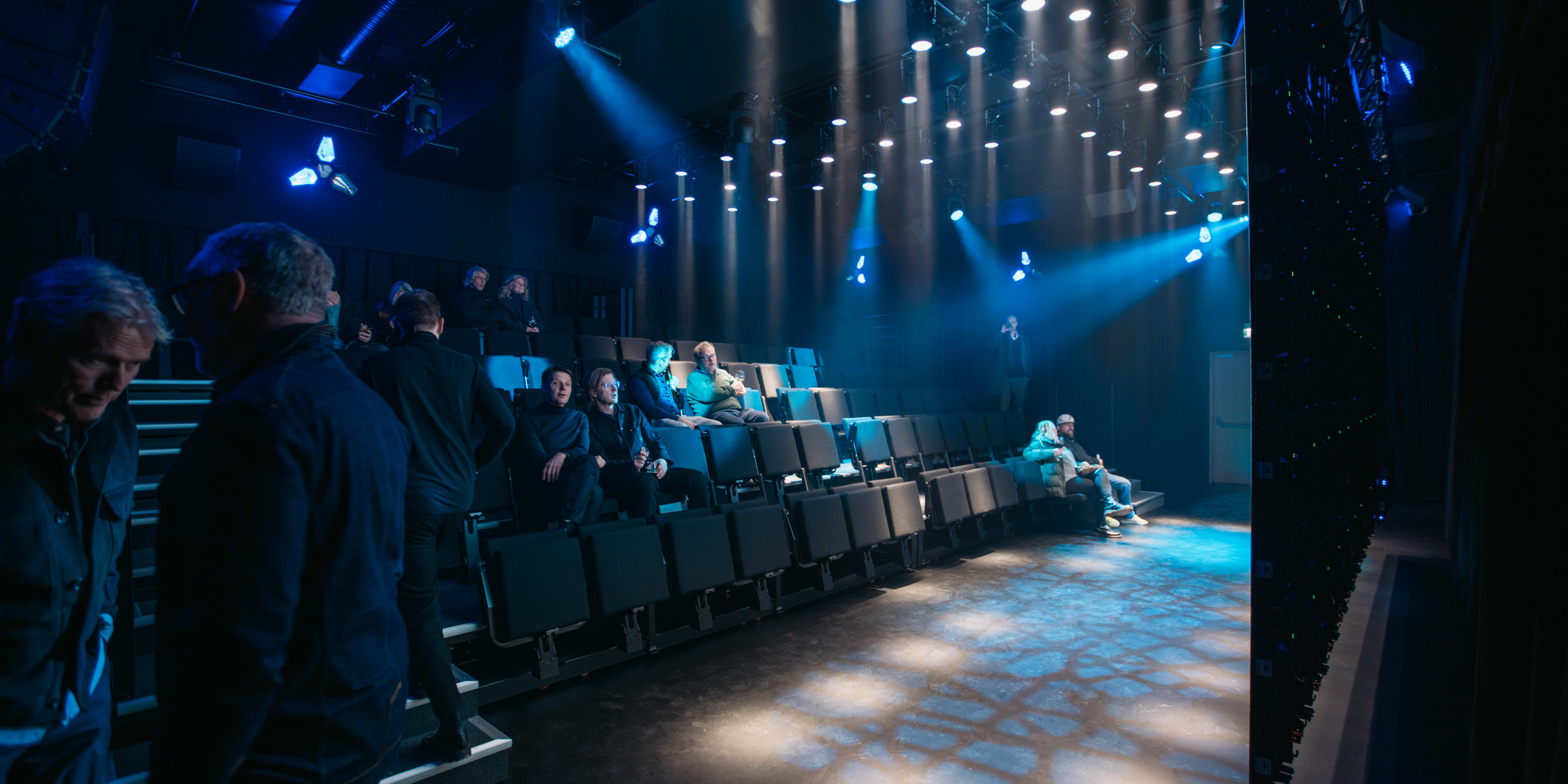 A group of people sitting in a dark theater with blue spotlights shining on them.  The people are wearing Norrøna clothing.  A large screen is behind them, with more blue lights on the ceiling above.