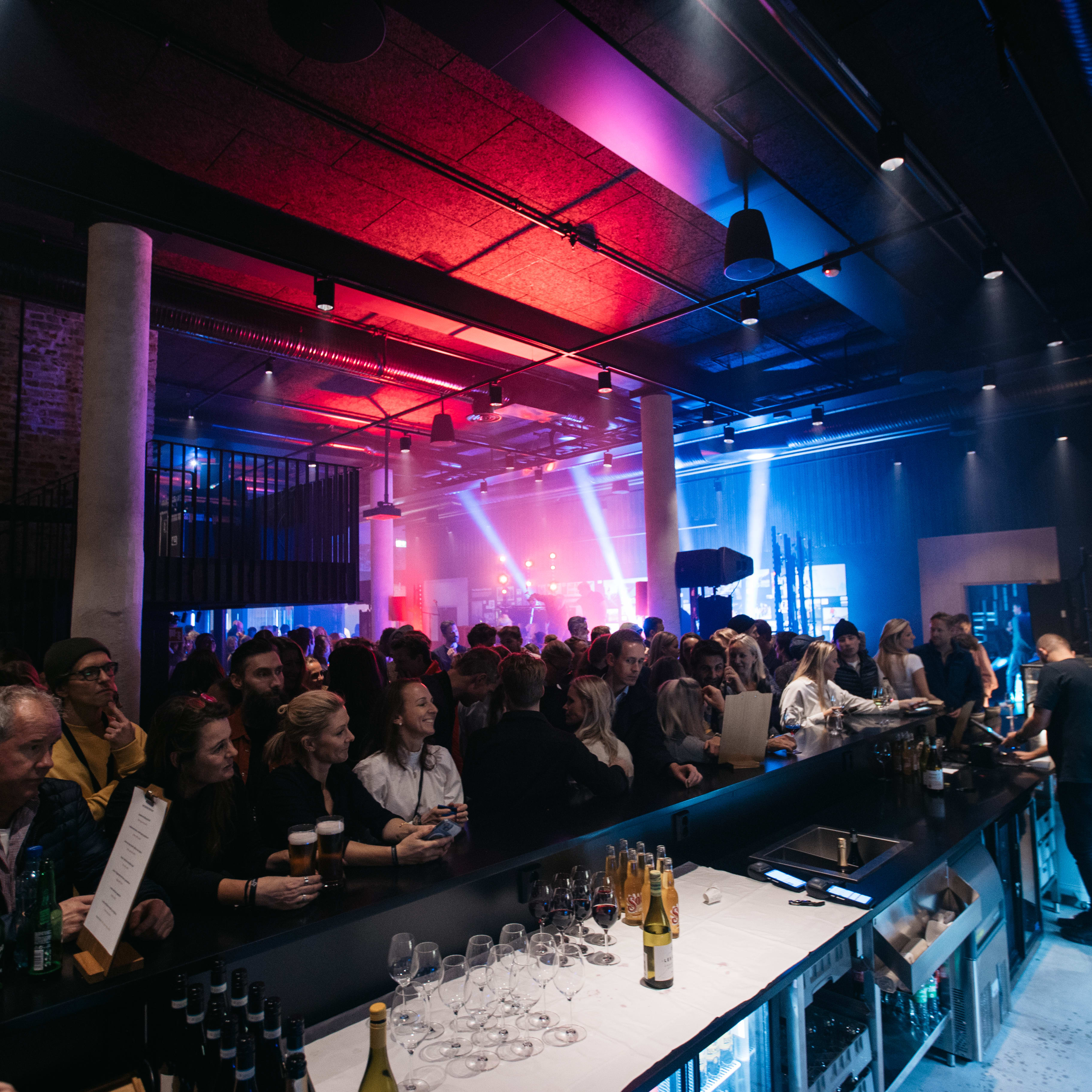 A group of people stand at a bar in a dimly lit room.  They are wearing Norrøna clothes and some are holding drinks.  There are several bottles of alcohol on the bar, as well as wine glasses and a few empty beer glasses.  The bar is lit with red and blue lights, casting shadows on the walls.  There are several other people standing in the background and a person to the right is working behind the bar.