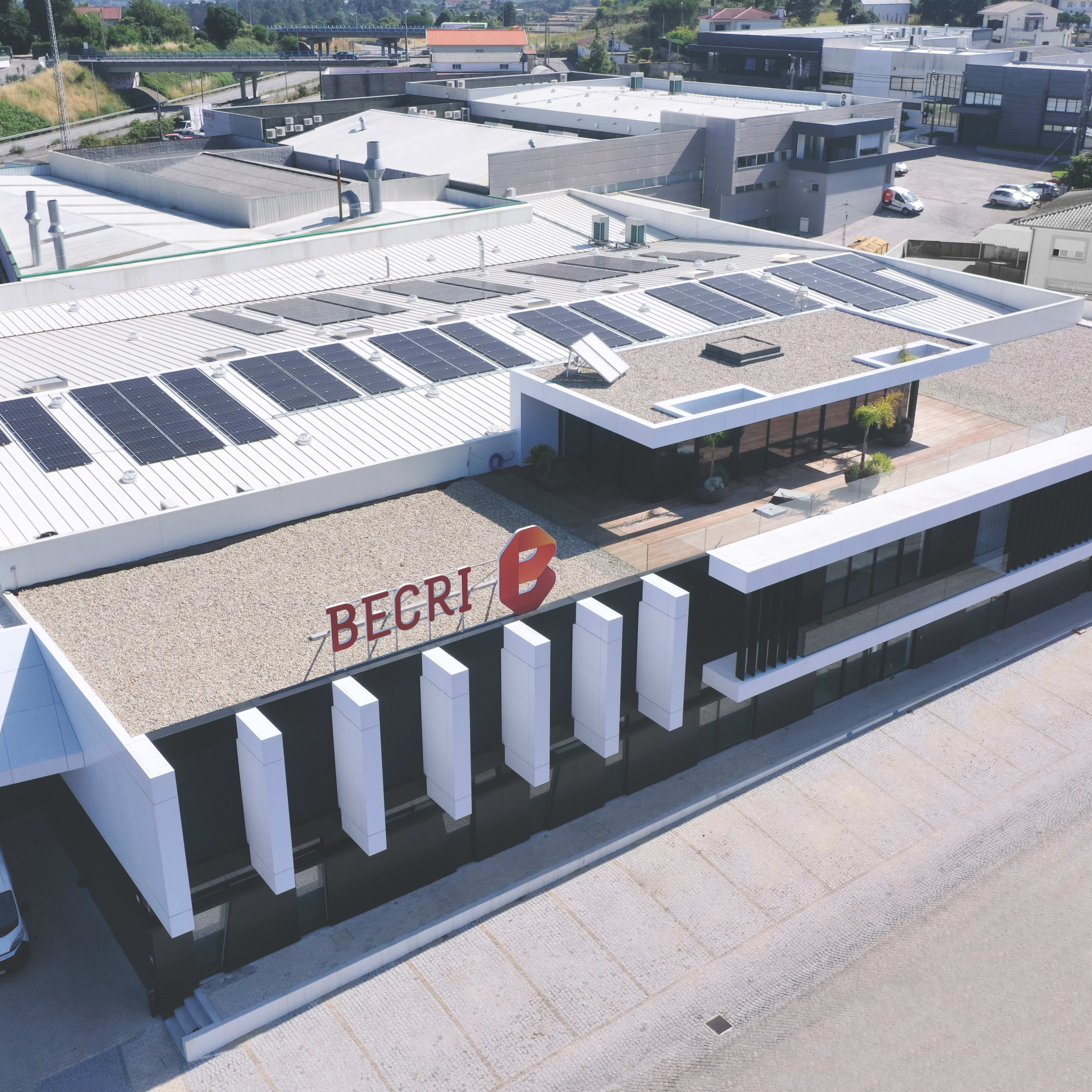 Aerial view of a building with a white roof and solar panels. The building has a sign that reads "BECRI". There is a white van parked in the parking lot.