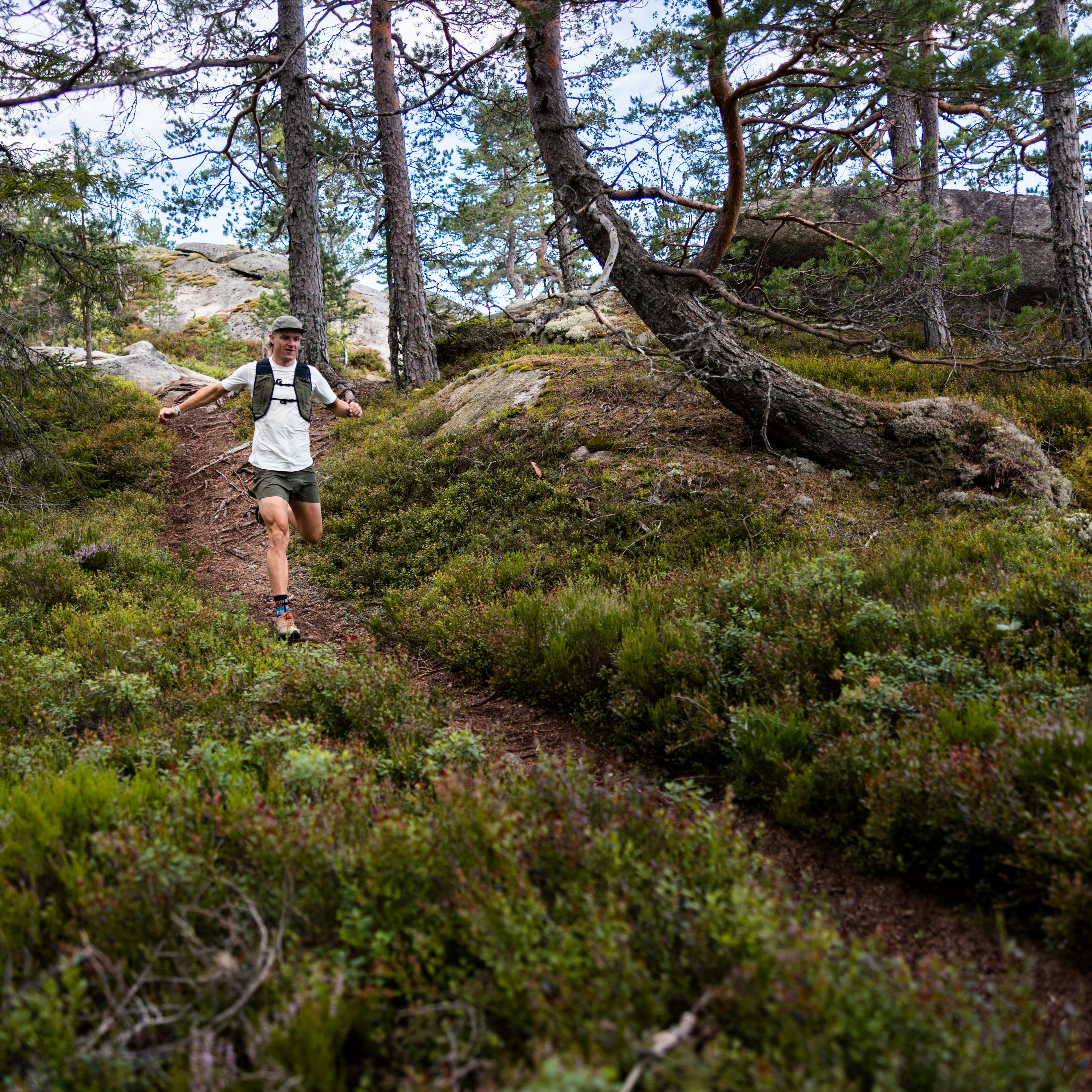 A man trail running downhill through a forest. He is wearing Norrøna shorts and a vest.
