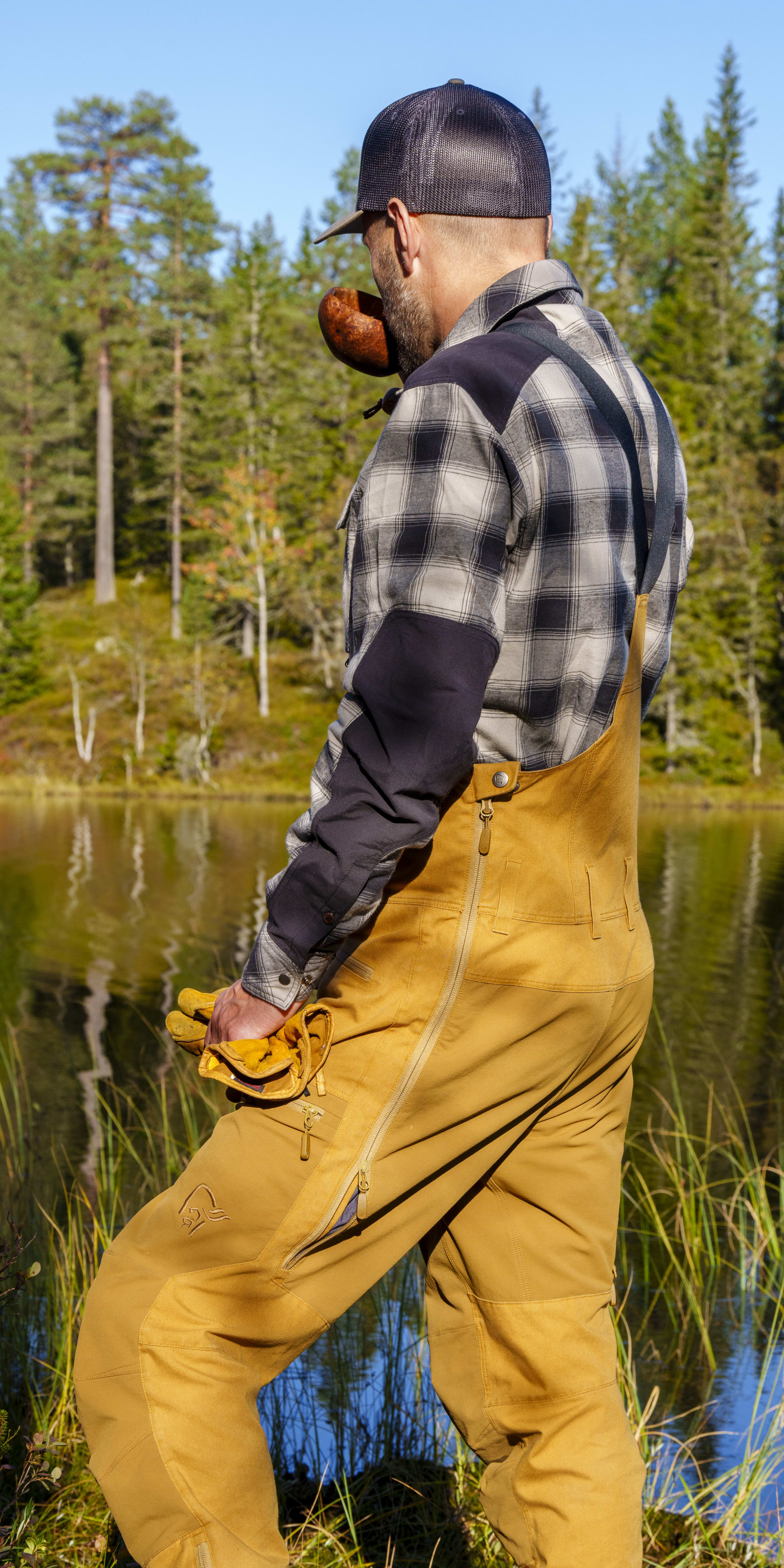 A man wearing a Norrøna  jacket stands by a lake in a forest. 
