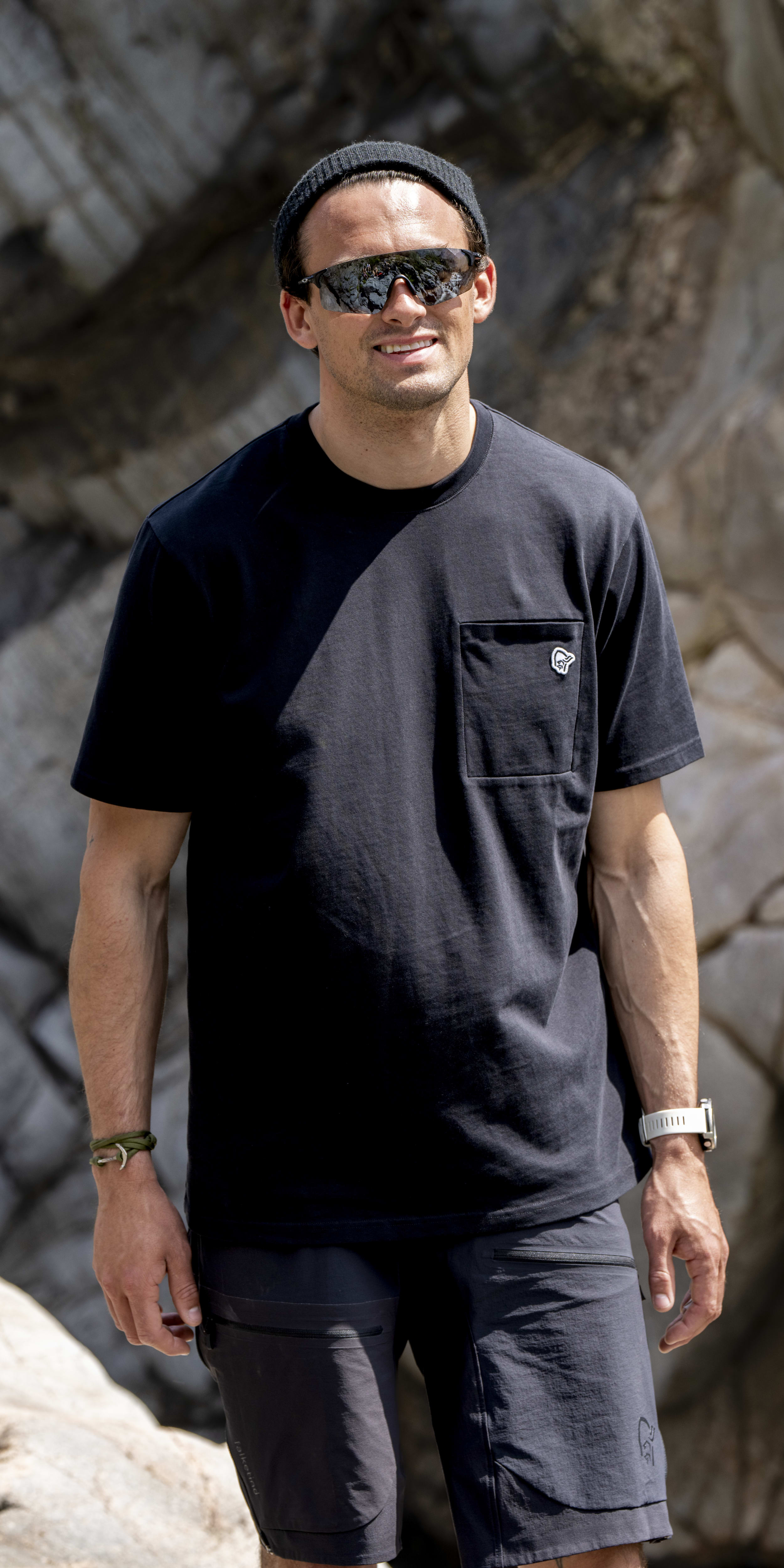 A man wearing a black Norrøna t-shirt and black Norrøna shorts, smiling and standing against a rocky backdrop. 
