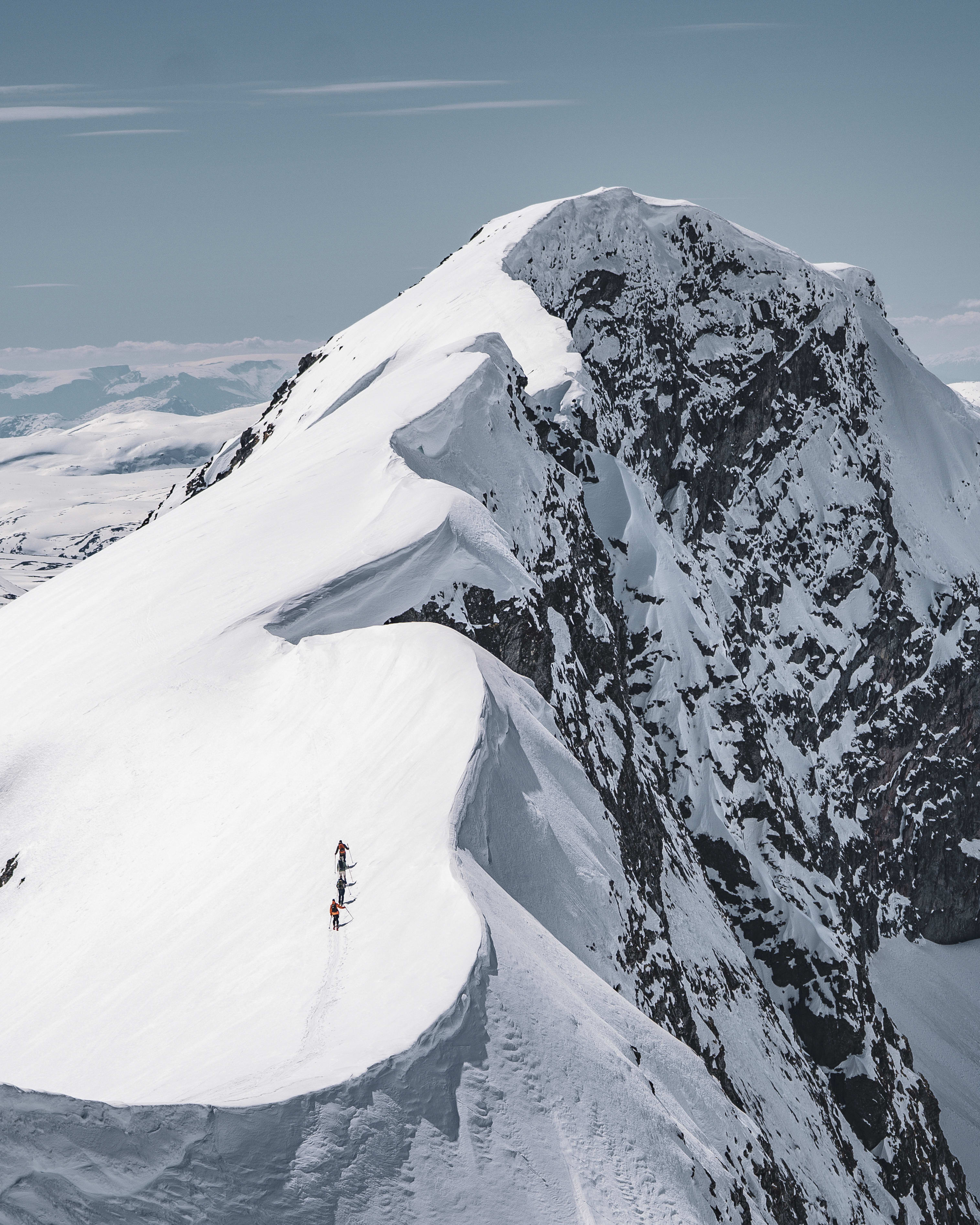 Three hikers ascend a snow-covered mountain ridge. The hikers wear bright orange jackets and are using ski poles for support.