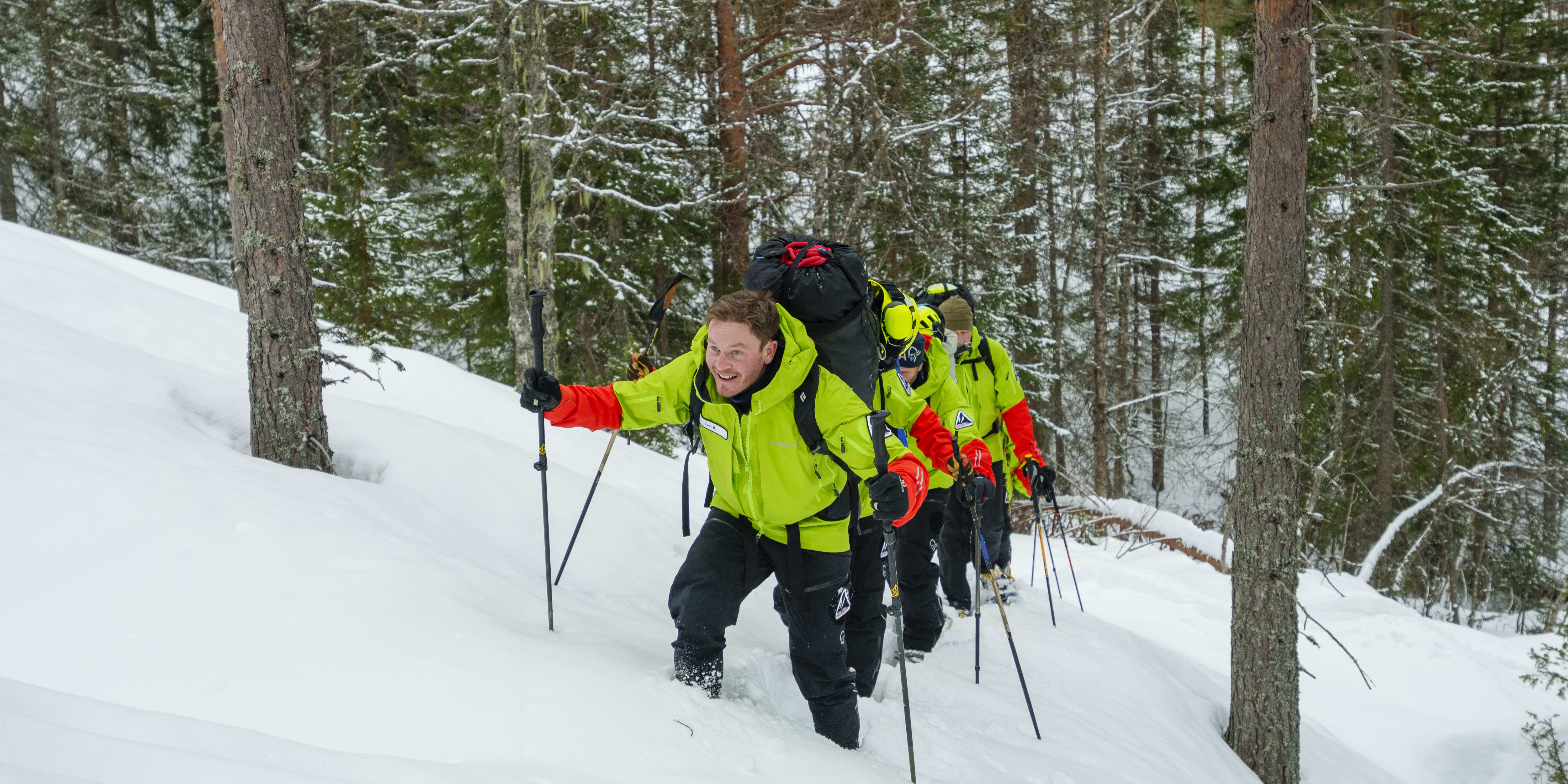 A man in a green Norrøna jacket walks through a snowy forest. He is smiling and using trekking poles. 
