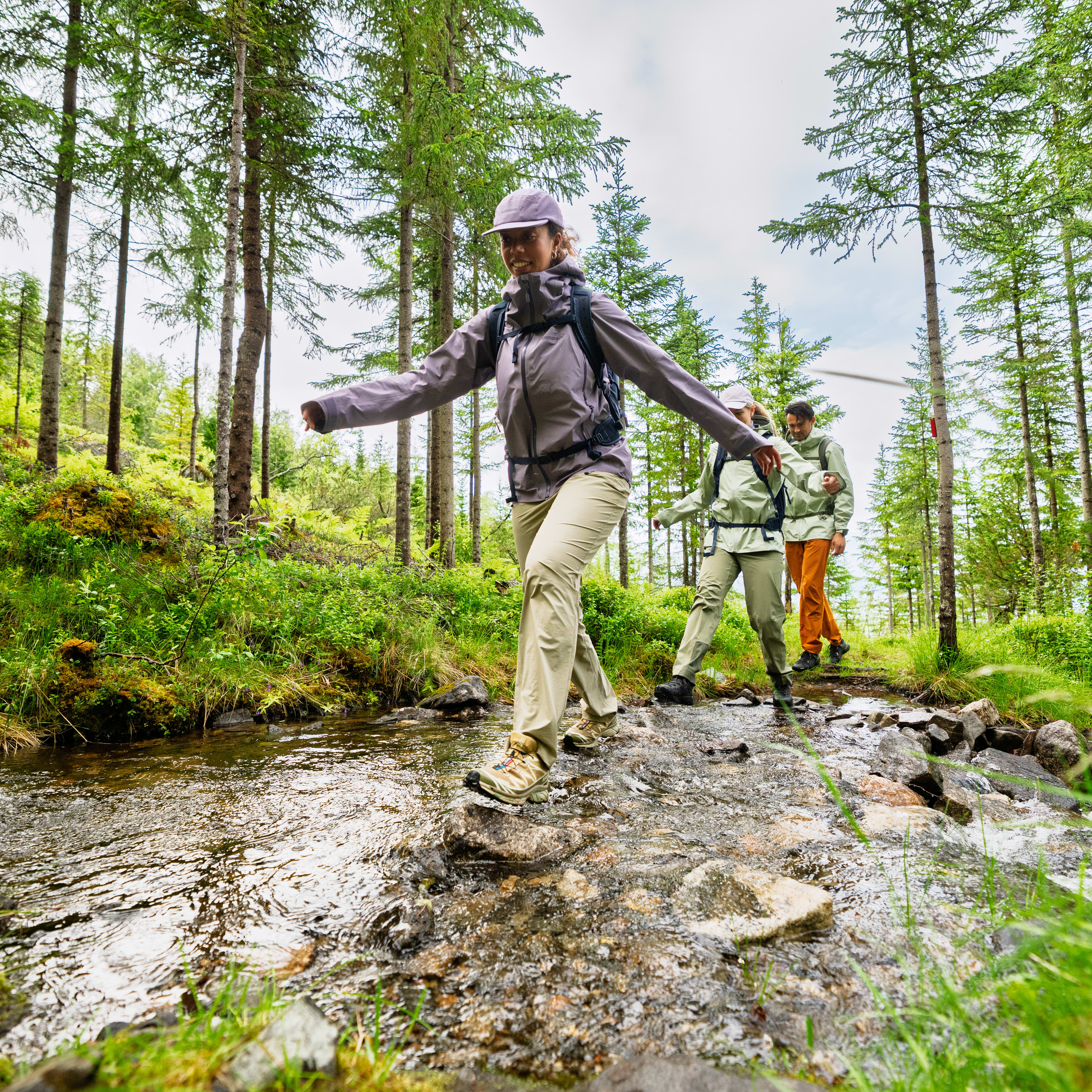 Three hikers in Norrøna clothing cross a rocky stream in a lush forest.