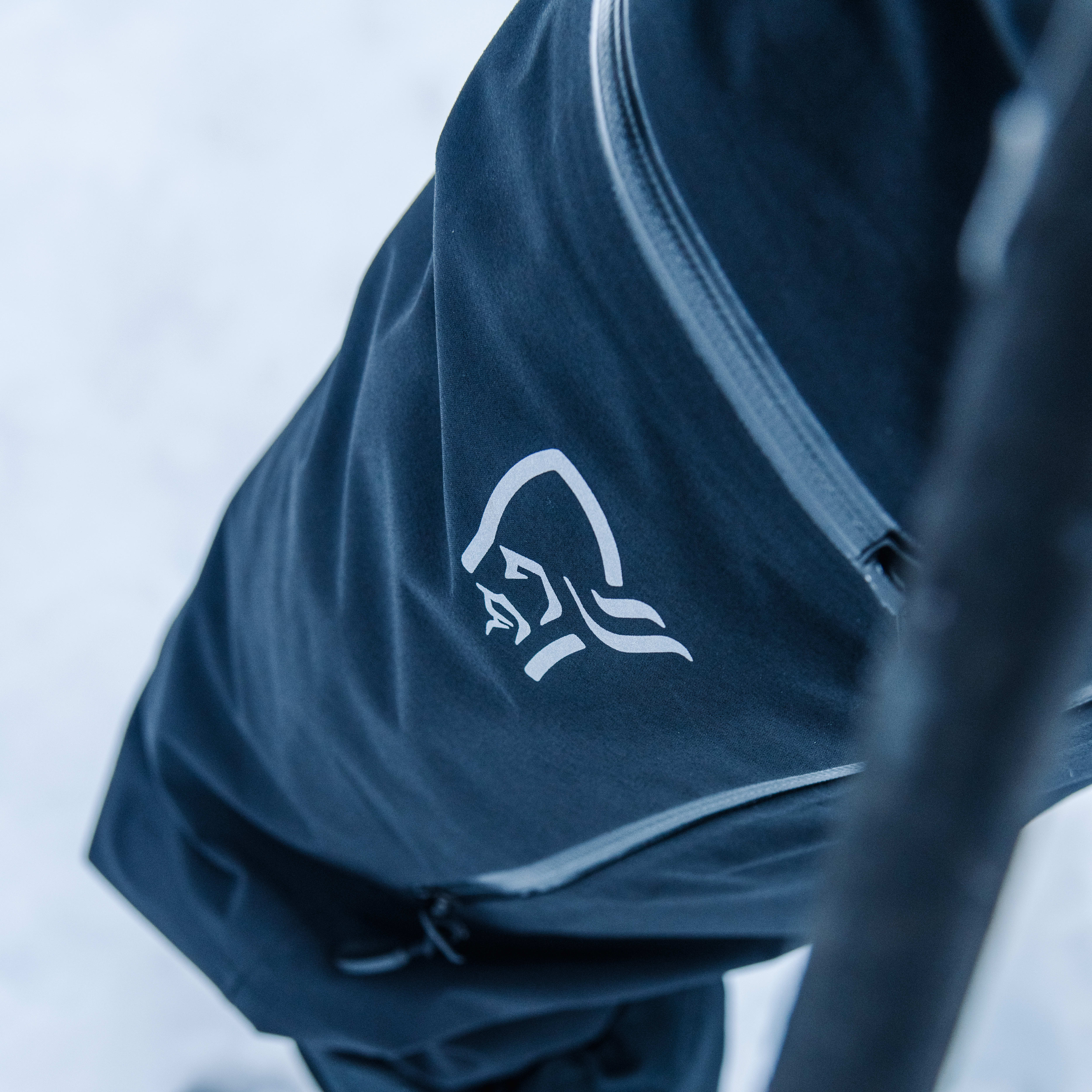 Closeup of a Norrøna pant leg with a white logo on a dark blue background.  A black trekking pole and yellow boot are out of focus in the background. 
