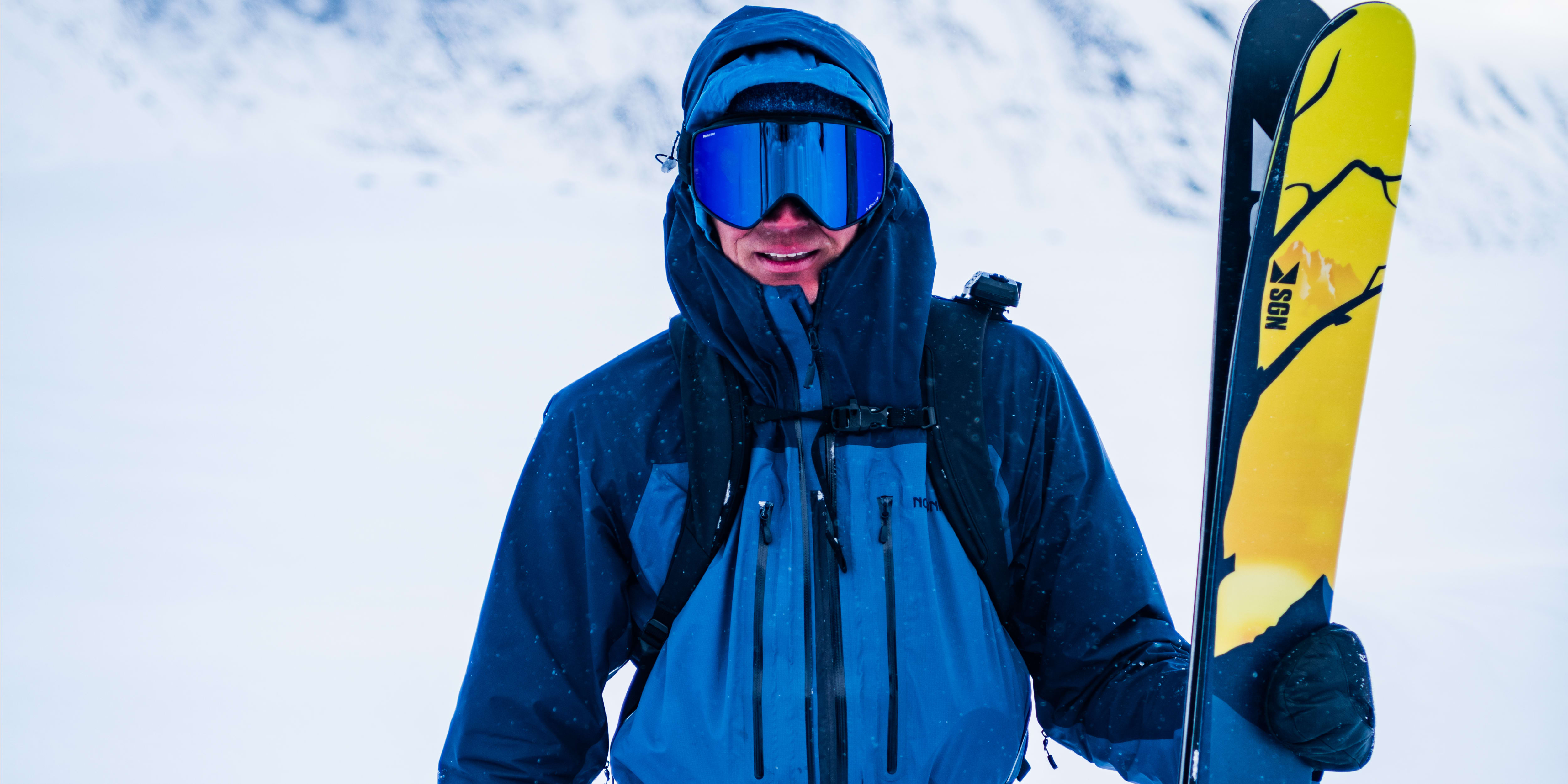 A man wearing Norrøna ski gear stands in snowy mountains holding skis.