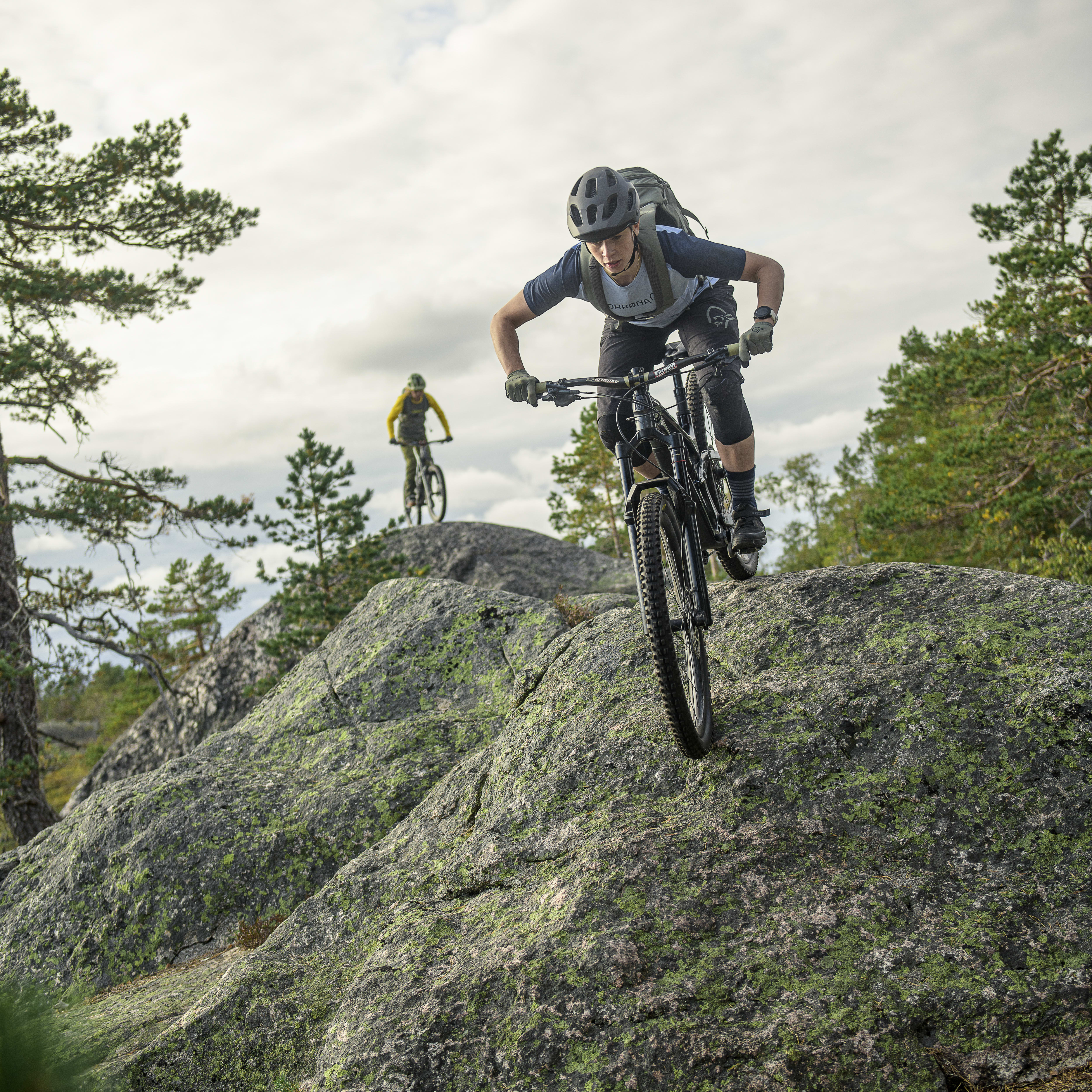 A mountain biker wearing Norrøna clothing rides down a rocky hill. Another biker follows behind. Trees and a cloudy sky fill the background.