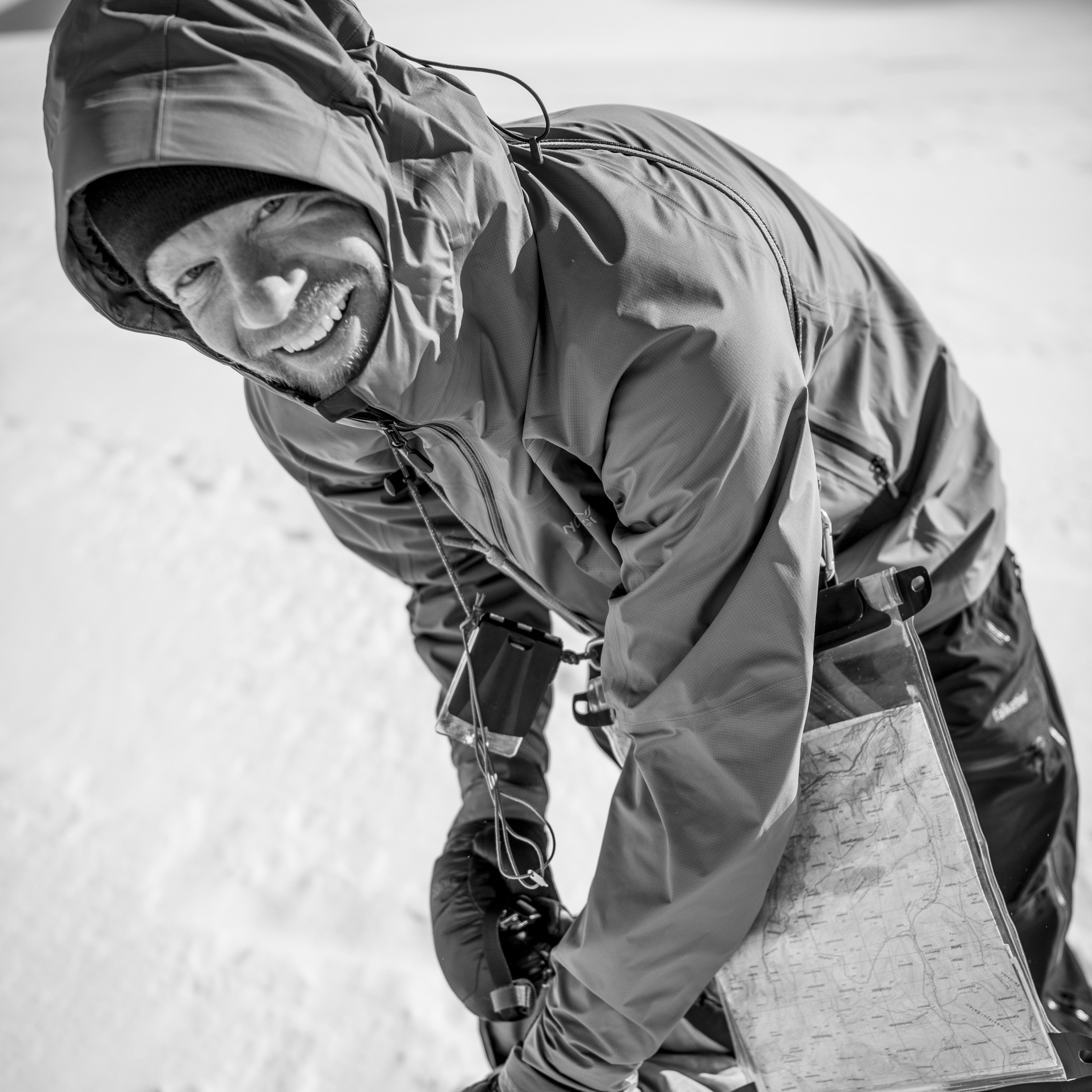 A man wearing a Norrøna jacket smiles as he looks at a map in a clear plastic case. 
