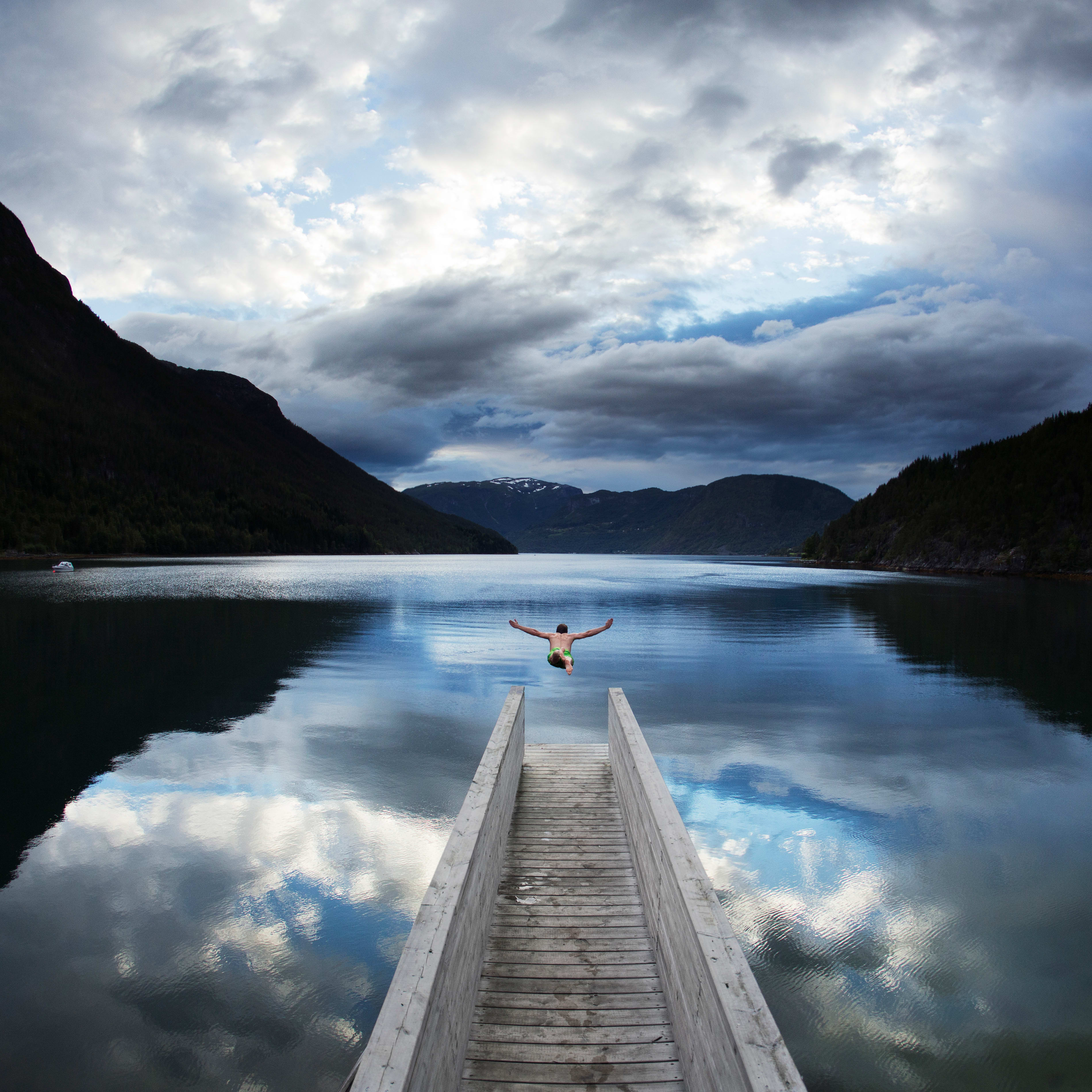 A person jumps off a wooden dock into a fjord. The sky above is partly cloudy, with the reflection of the clouds visible in the water.