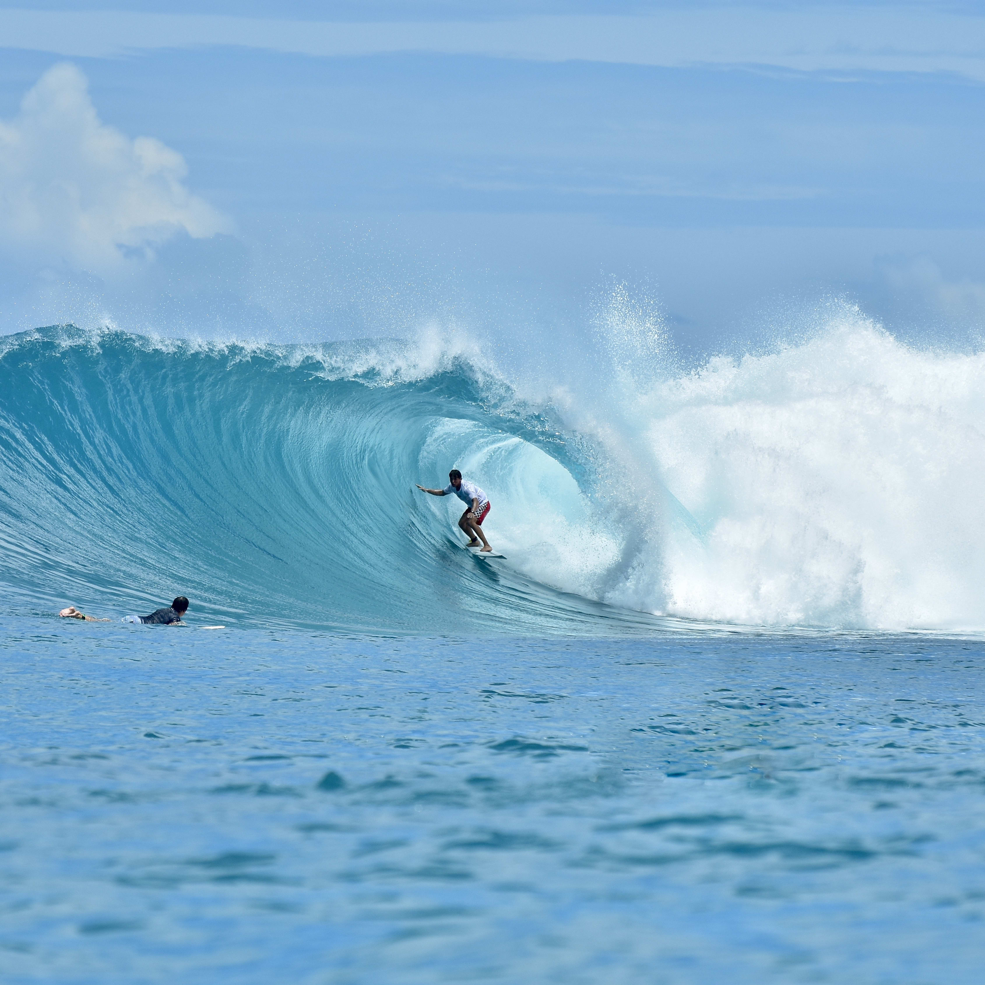 A surfer wearing Norrøna clothing carves through a large blue wave, with another person on a board in the foreground.