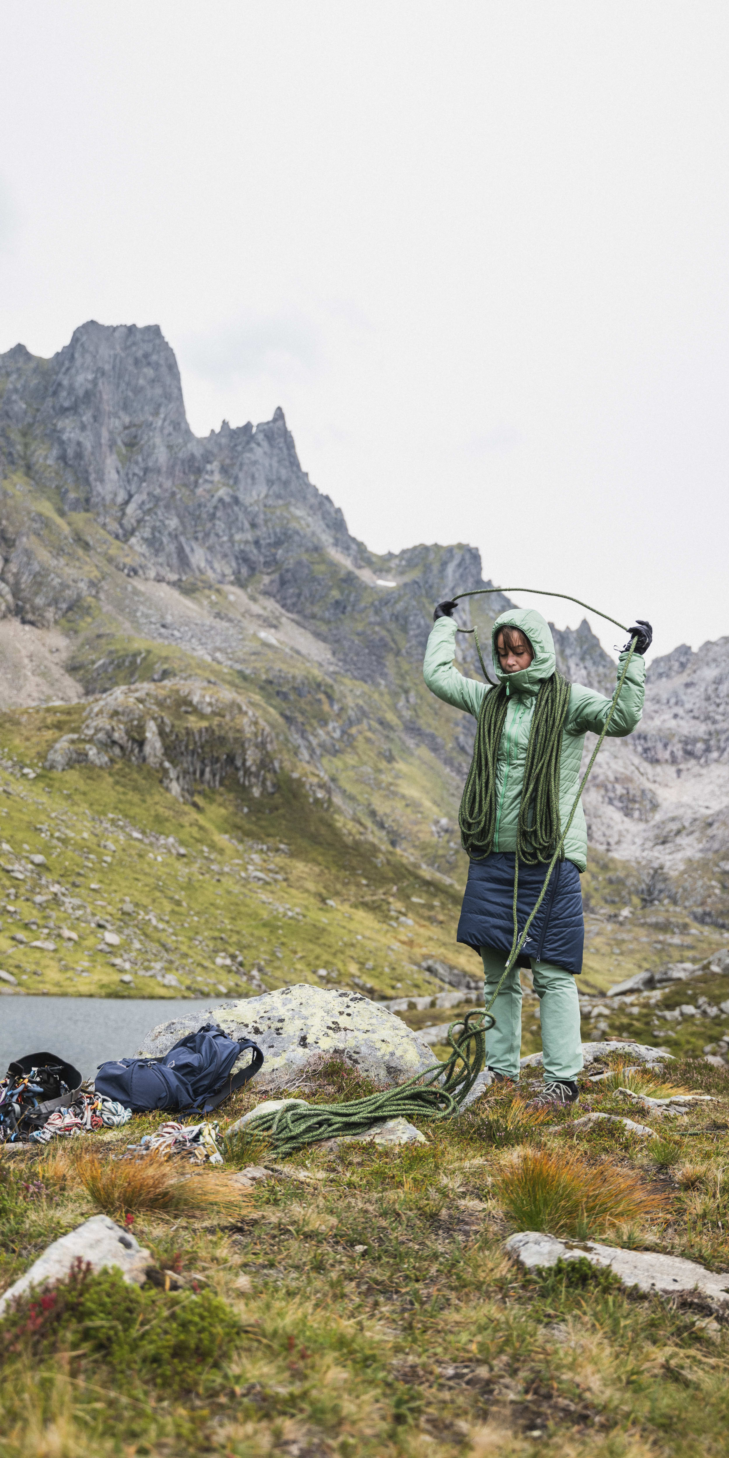 A woman wearing a Norrøna jacket and pants holds a climbing rope in front of a mountain lake. 
