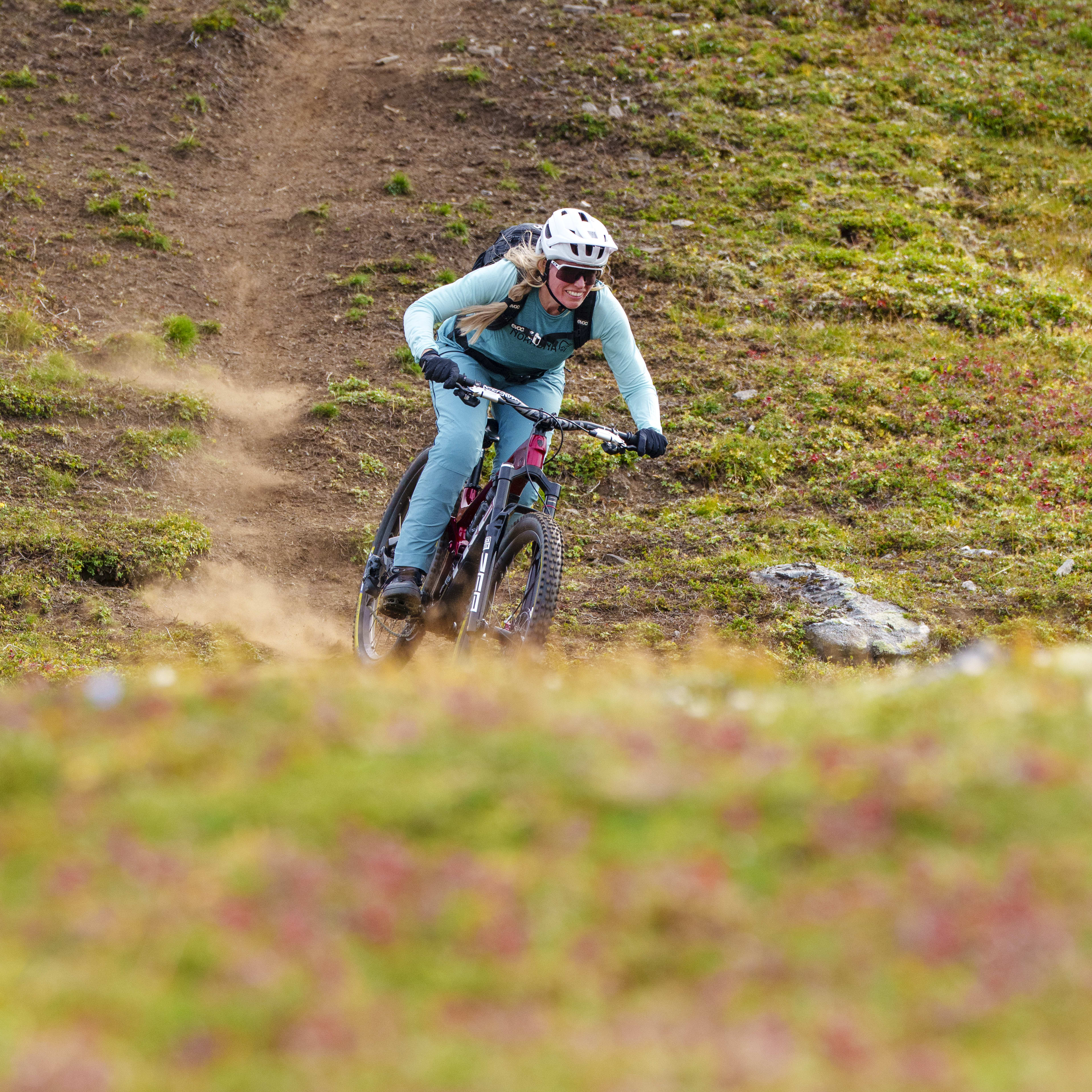 A woman mountain bikes on a dirt trail in a vibrant, green, and red meadow. She wears a Norrøna turquoise outfit and helmet.

