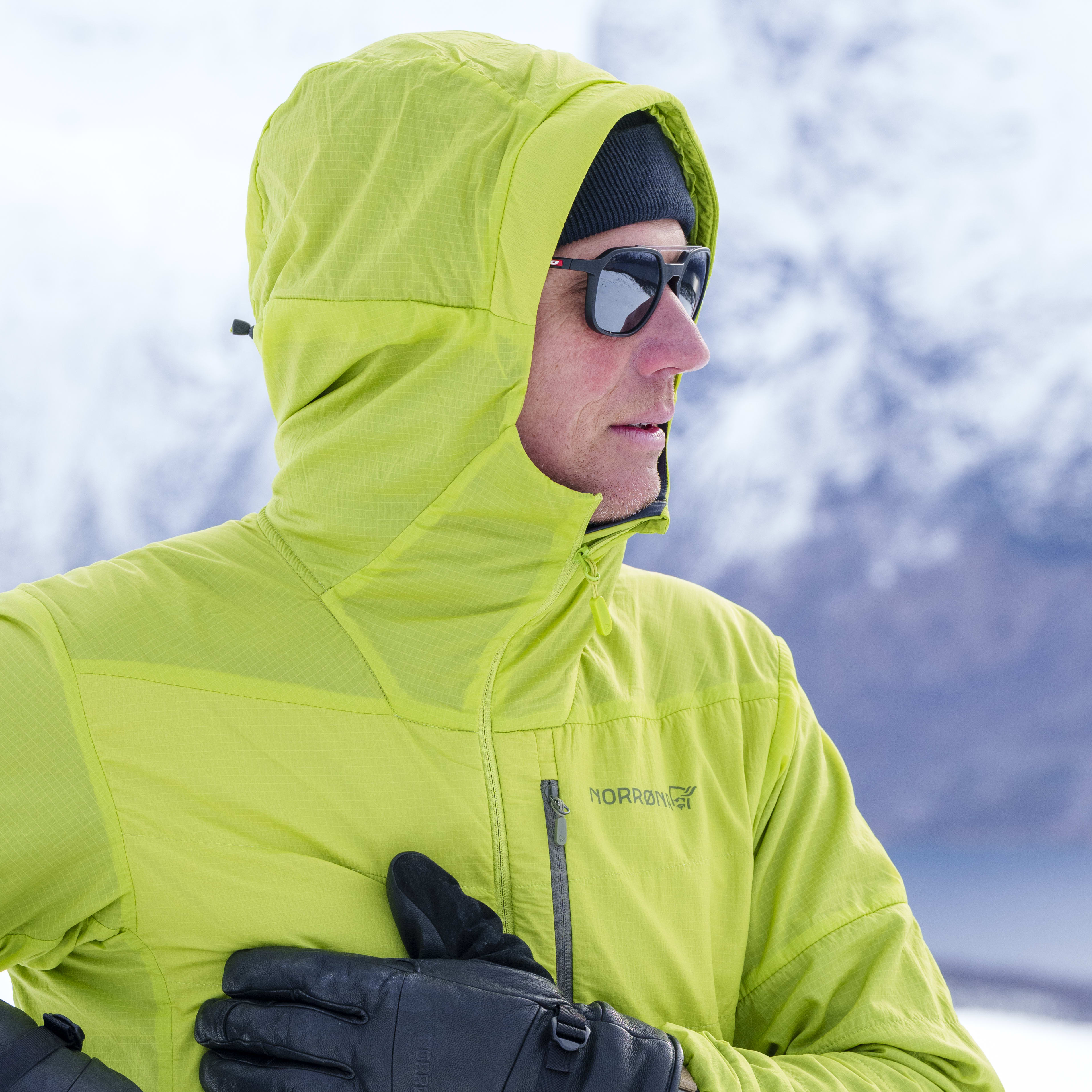 Man in sunglasses wearing a hooded Norrøna jacket and gloves stands outdoors in a snowy mountain landscape.