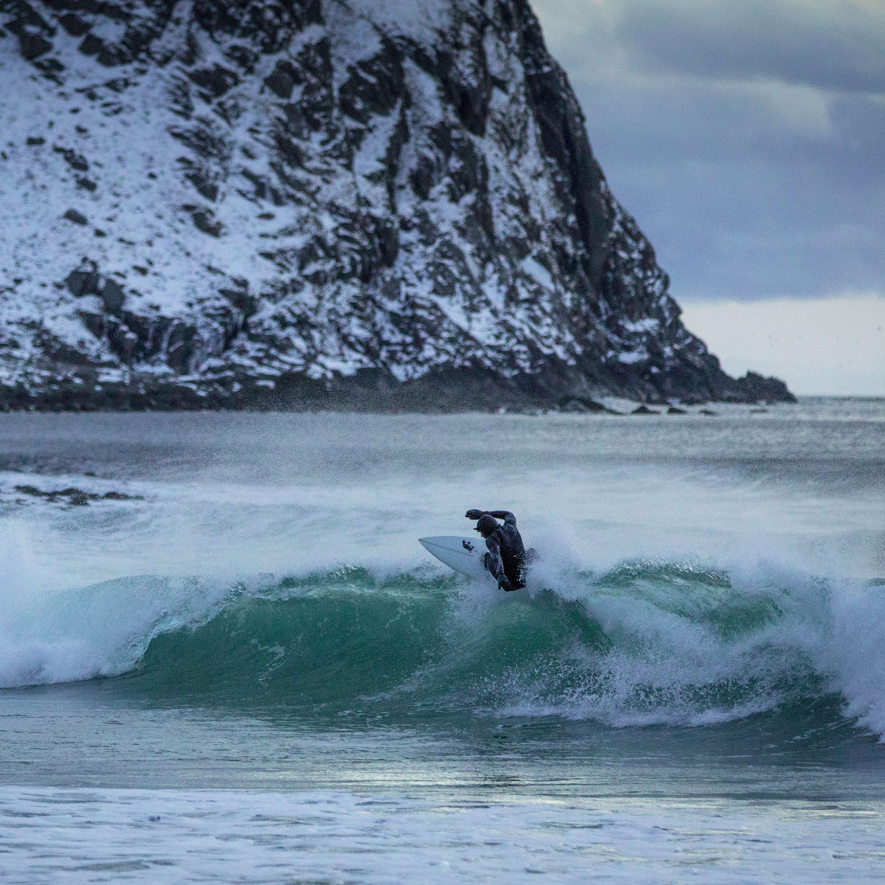 A surfer, wearing a Norrøna wetsuit, rides a turquoise wave near a snow-covered mountain under a cloudy sky.