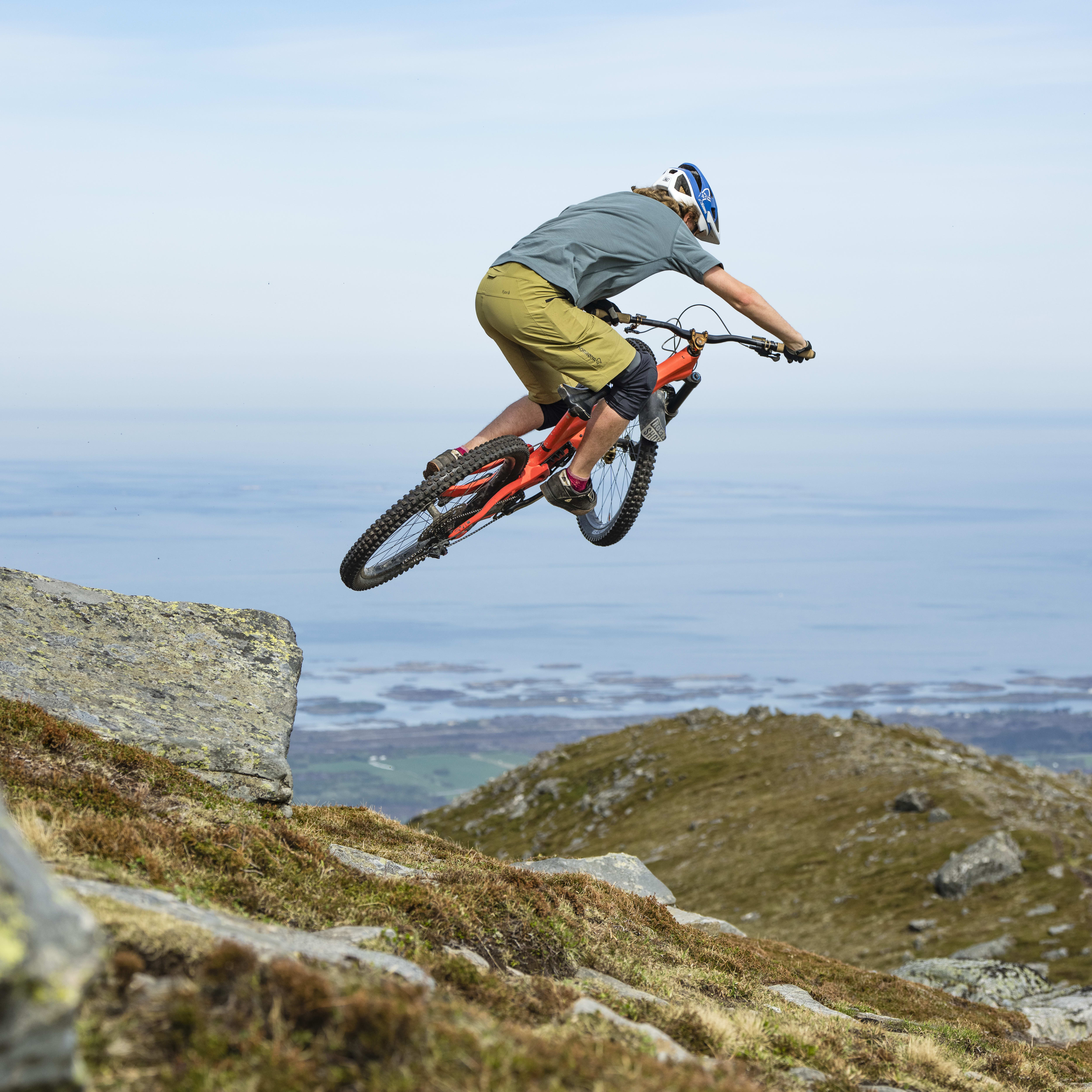 A person is mountain biking off a cliff and is in mid-air. The bike has a red frame and black tires. The person is wearing a helmet and knee pads. The person is going over a cliff edge that drops to a body of water in the distance. The background is blurred, and it appears the person is on a grassy mountain top.