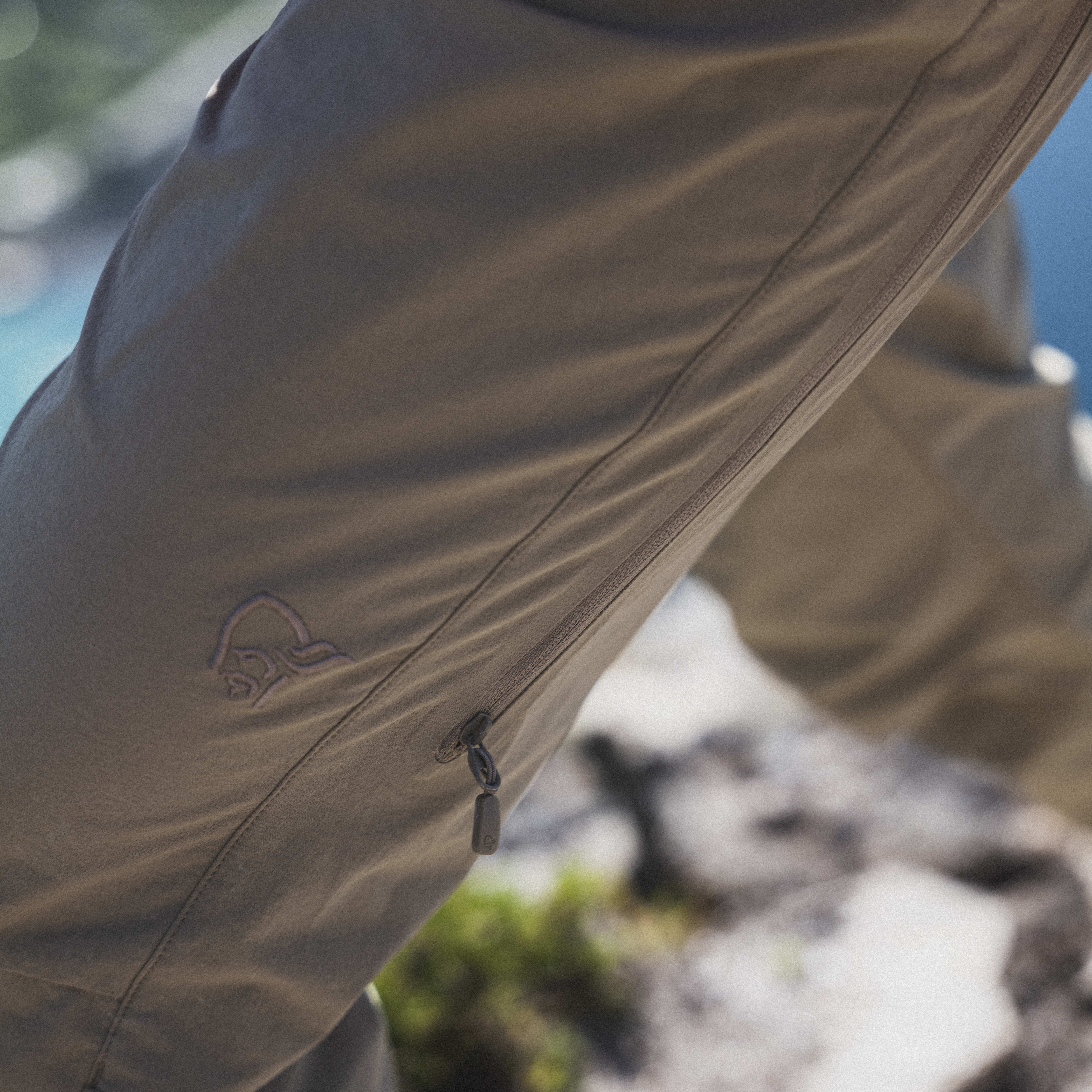 Close-up of a person wearing khaki Norrøna trekking pants, with a zipper detail and logo, against a blurred mountain landscape.