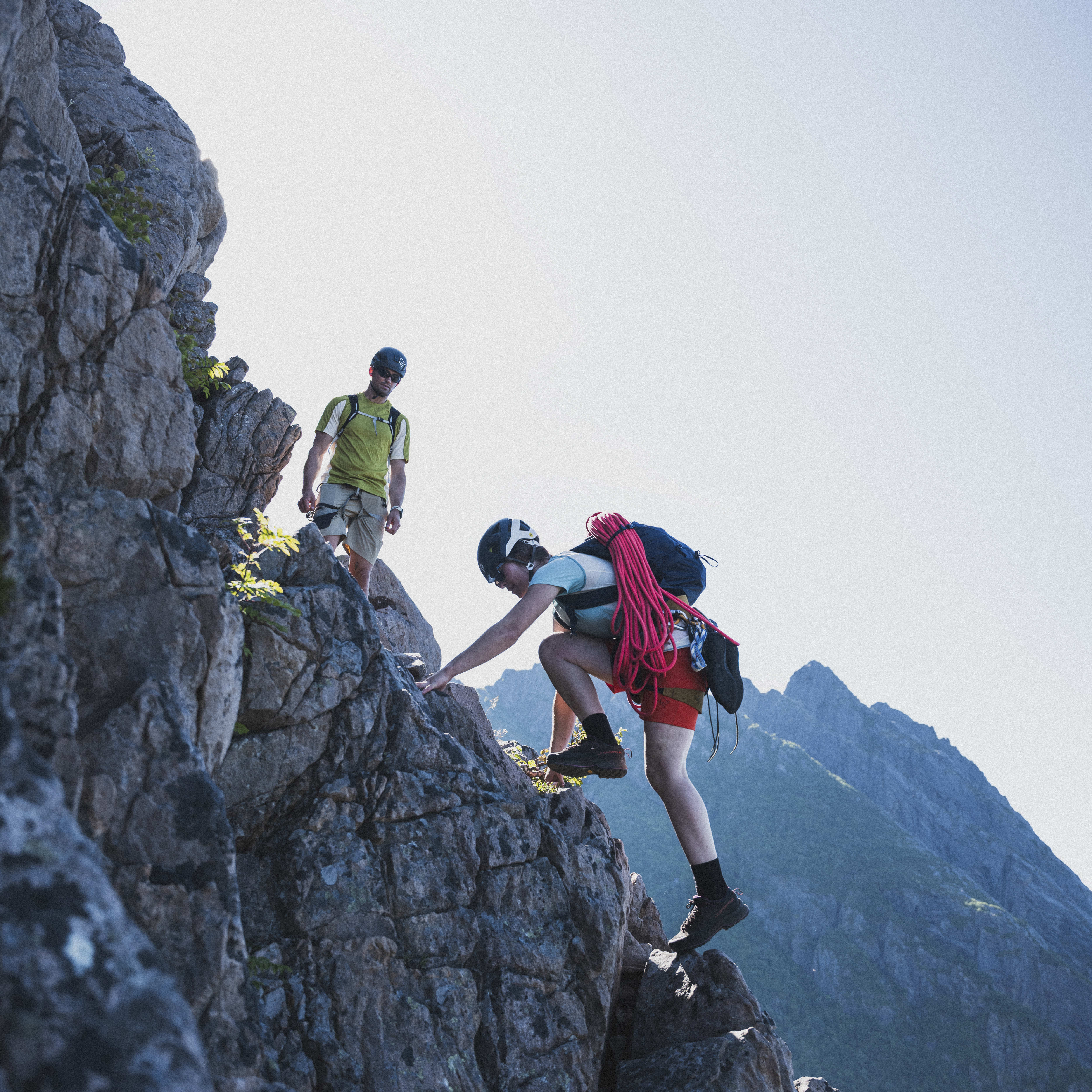Two climbers navigate a rocky cliff face against a clear sky and mountain backdrop. The lead climber, wearing a helmet, a Norrøna backpack and red shorts, ascends.