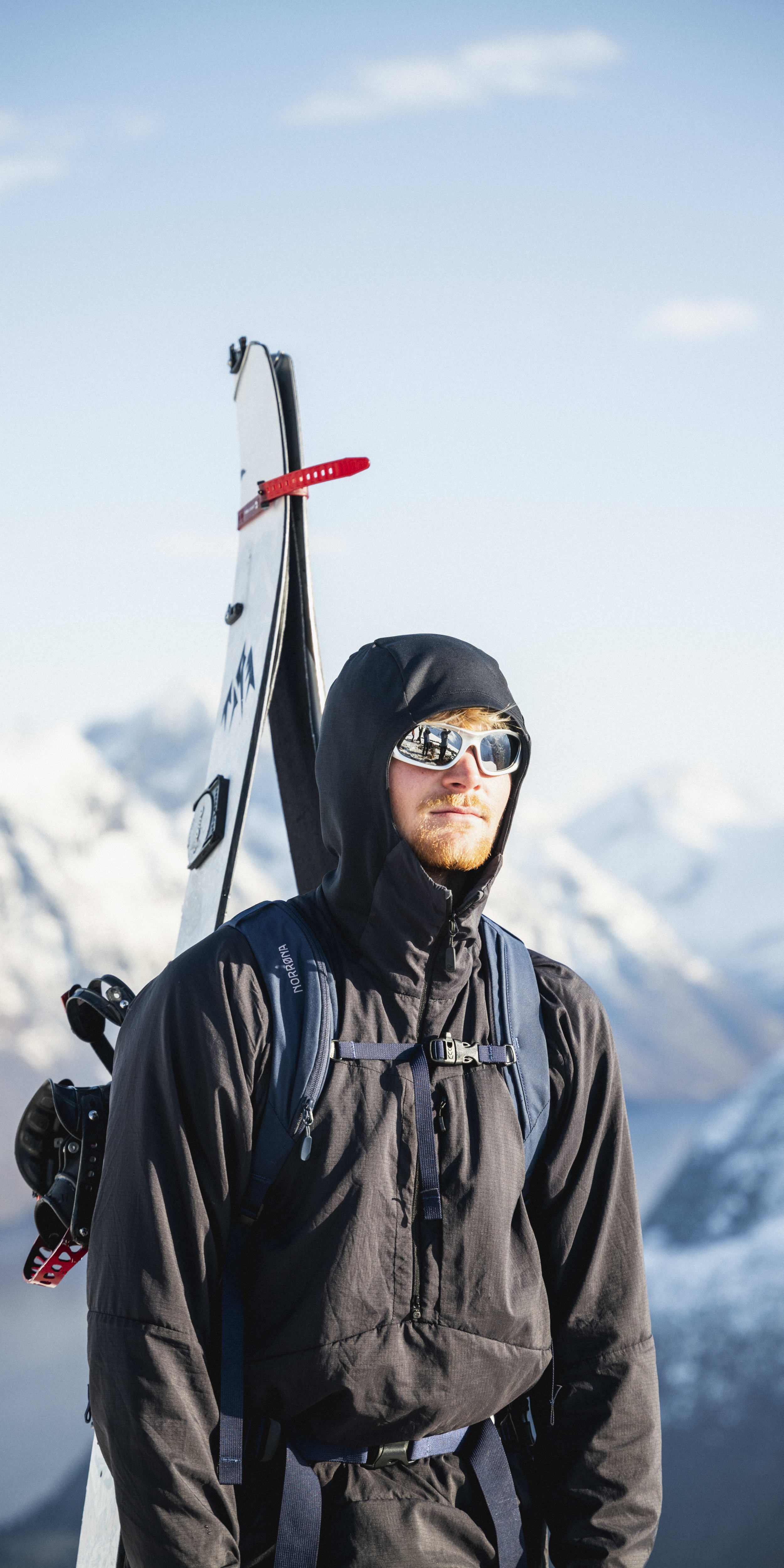 A man wearing a Norrøna jacket and sunglasses standing with a ski in his back, overlooking snowy mountains. 
