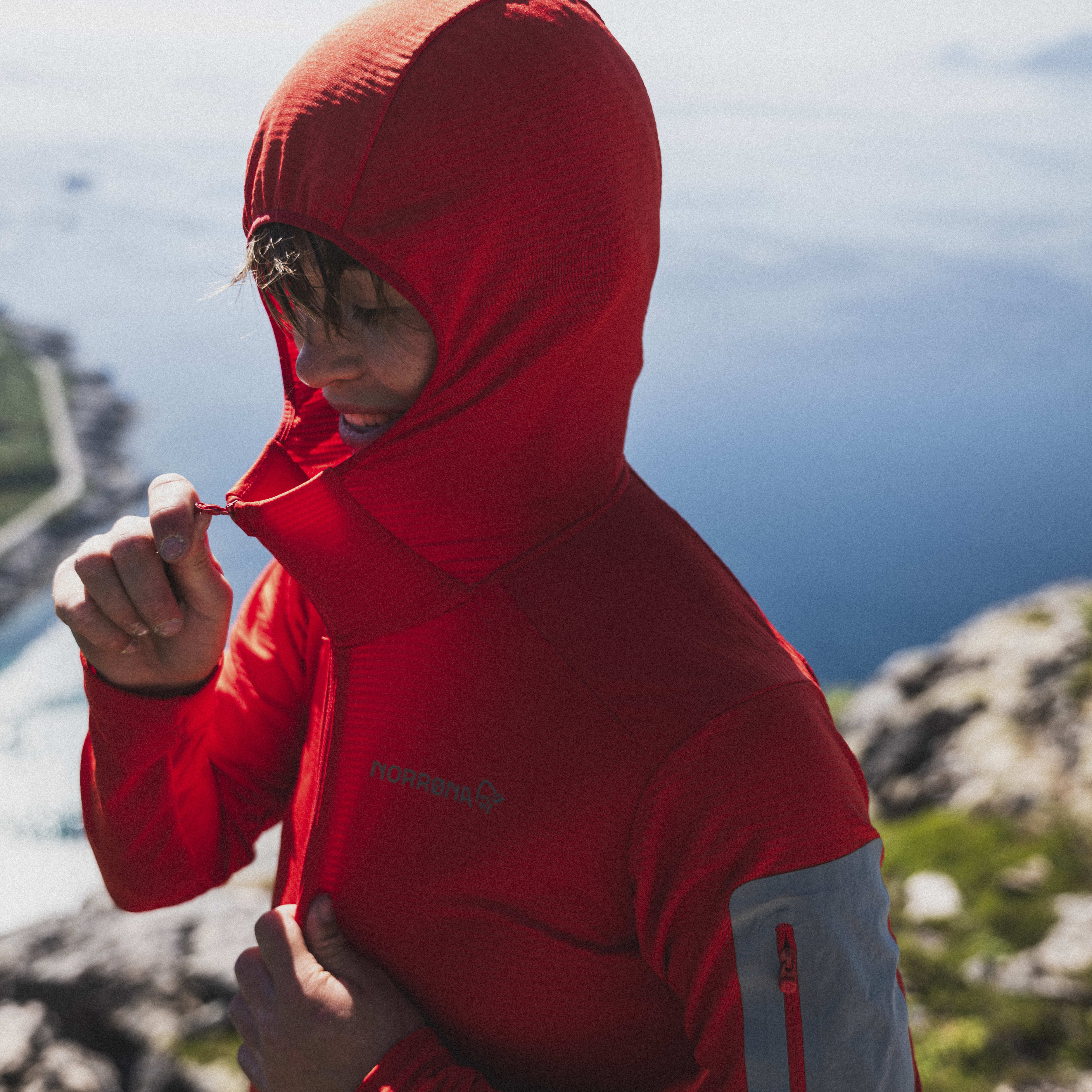 A person wearing a red Norrøna hooded fleece jacket adjusting the hood while on a mountain, with ocean scenery visible in the background.