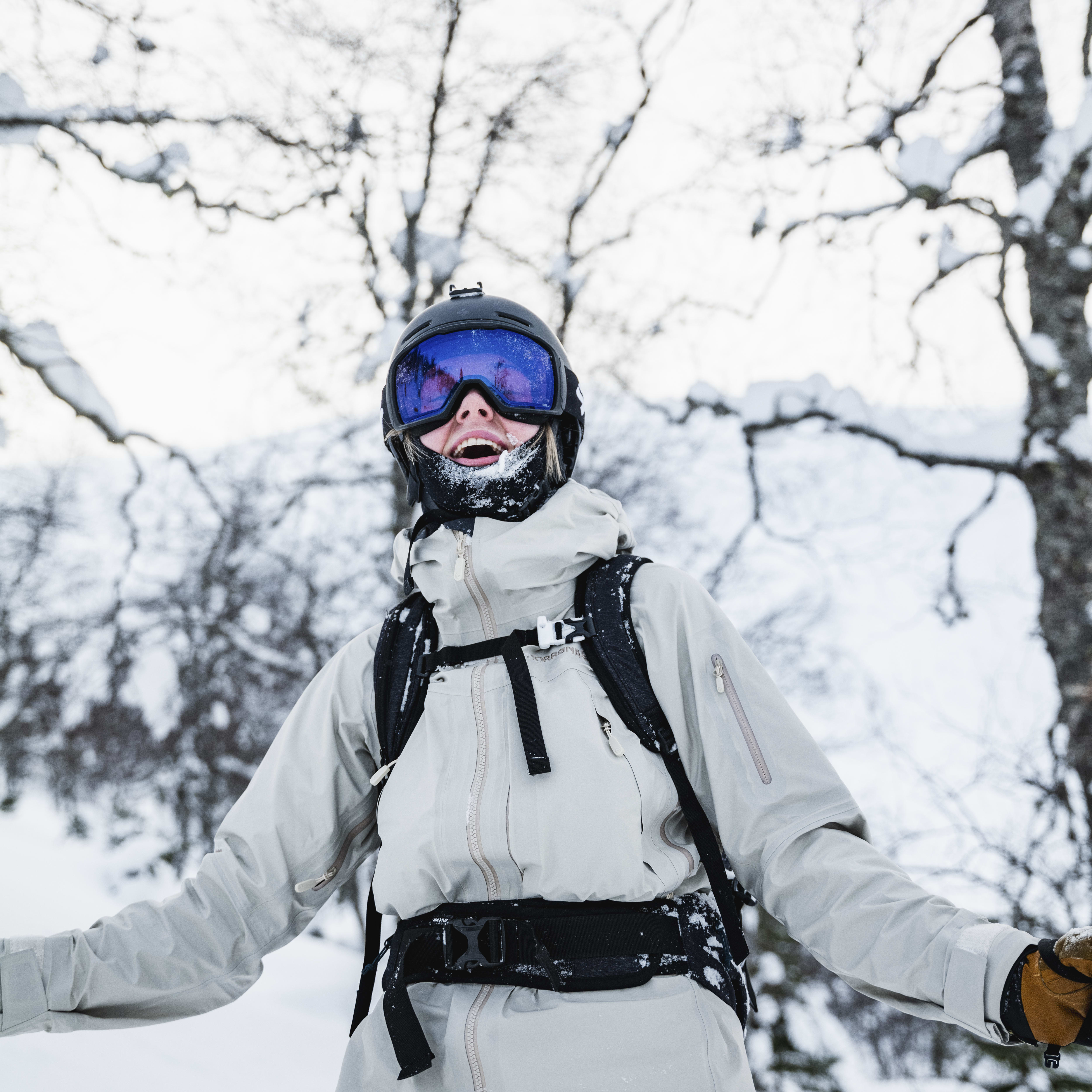 A person wearing a Norrøna ski jacket and pants smiles for the camera in a snowy forest. 
