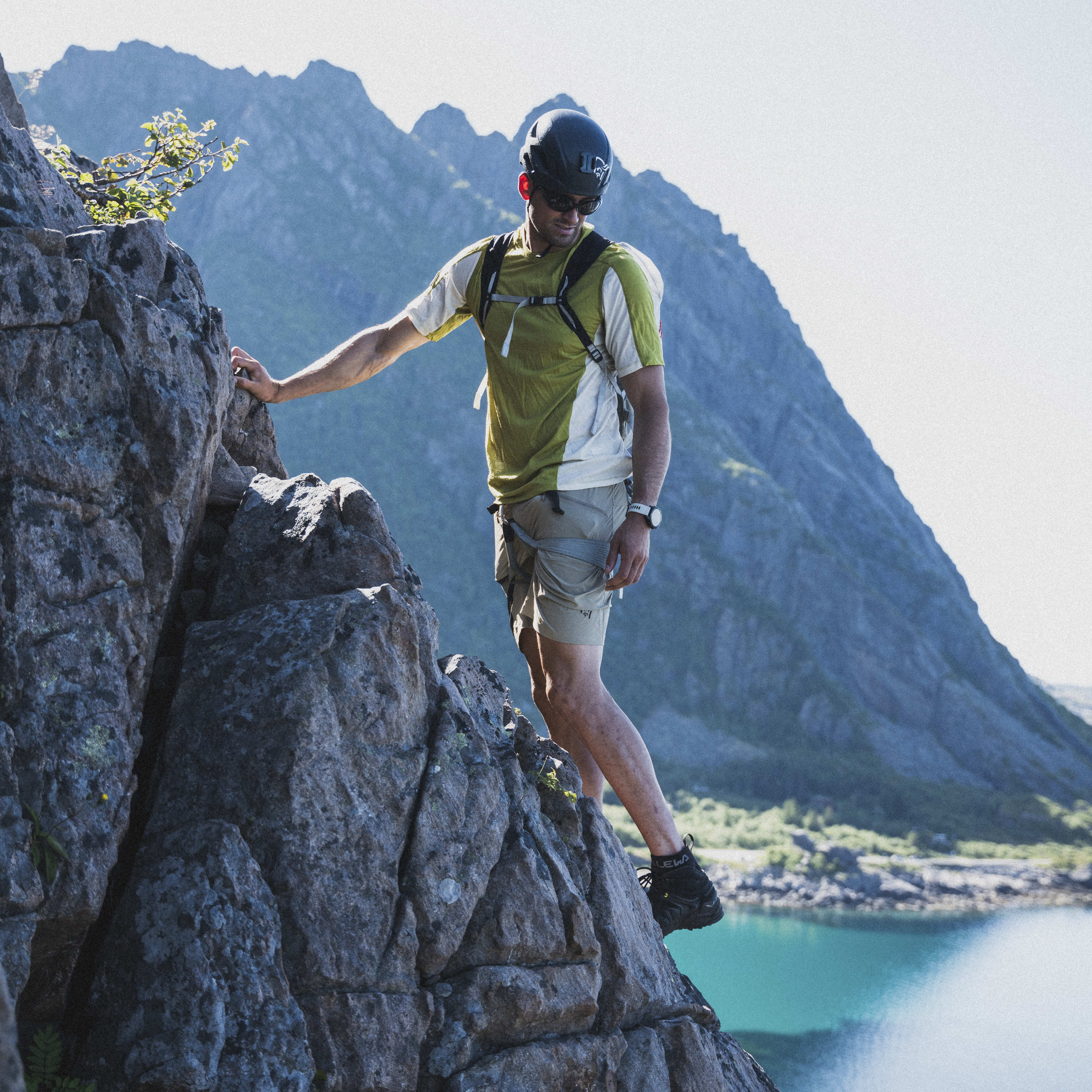 A climber wearing a Norrøna shirt, helmet, and backpack scales a rock face with a blue fjord and mountains in the background.
