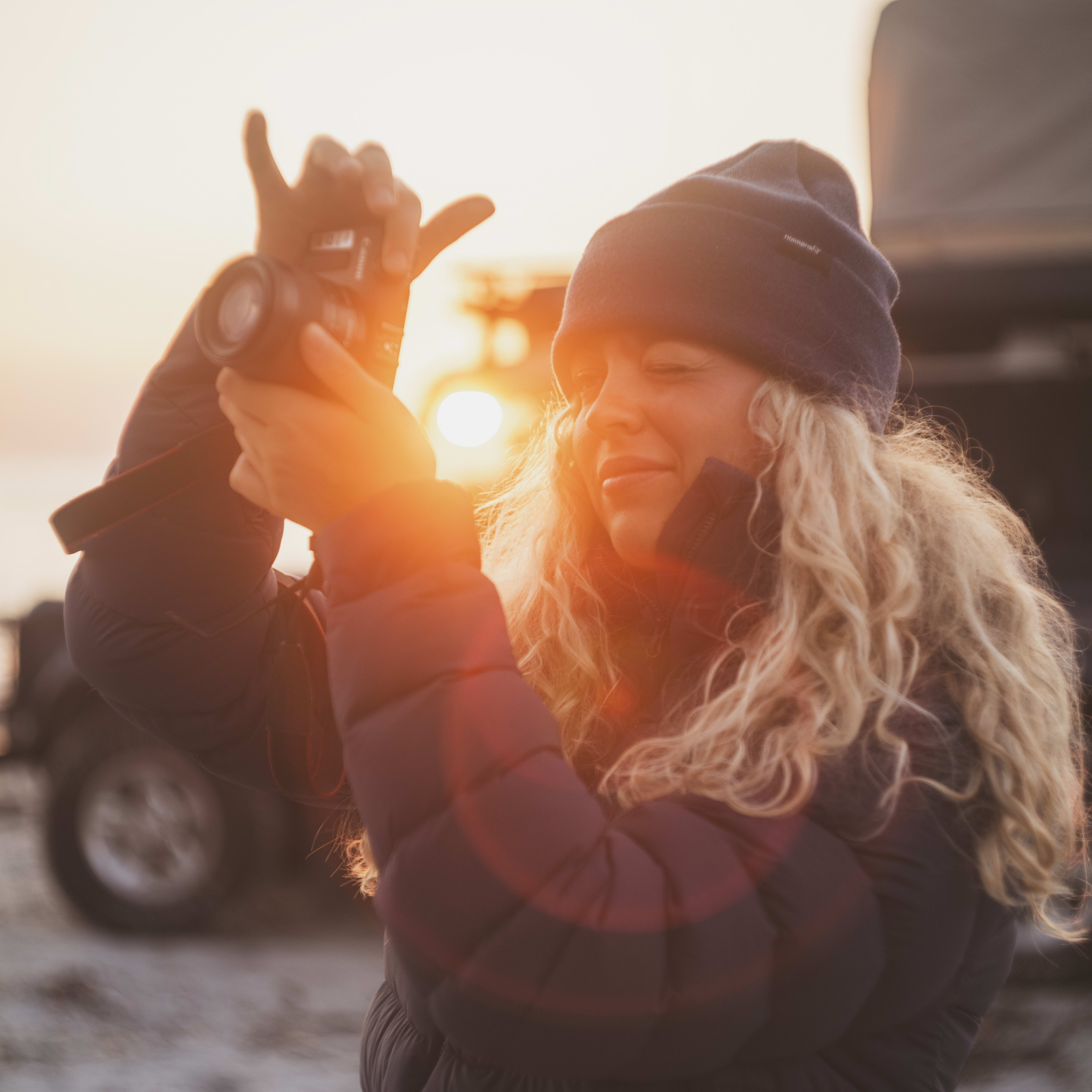 Woman in Norrøna jacket/beanie photographs a sunset. A car with a roof tent is behind.