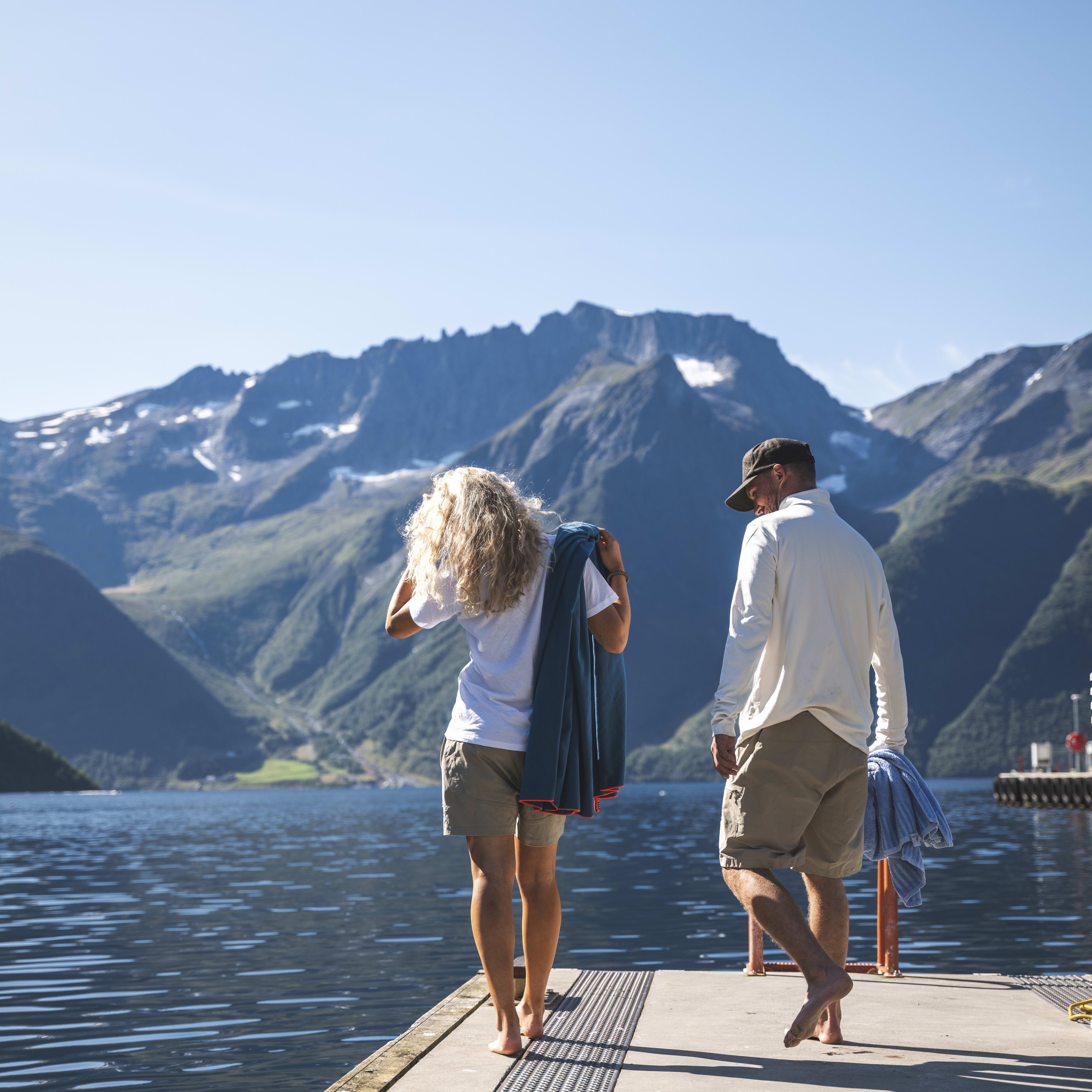 Couple walking on a dock toward mountains. They wear Norrøna shorts and carry towels.