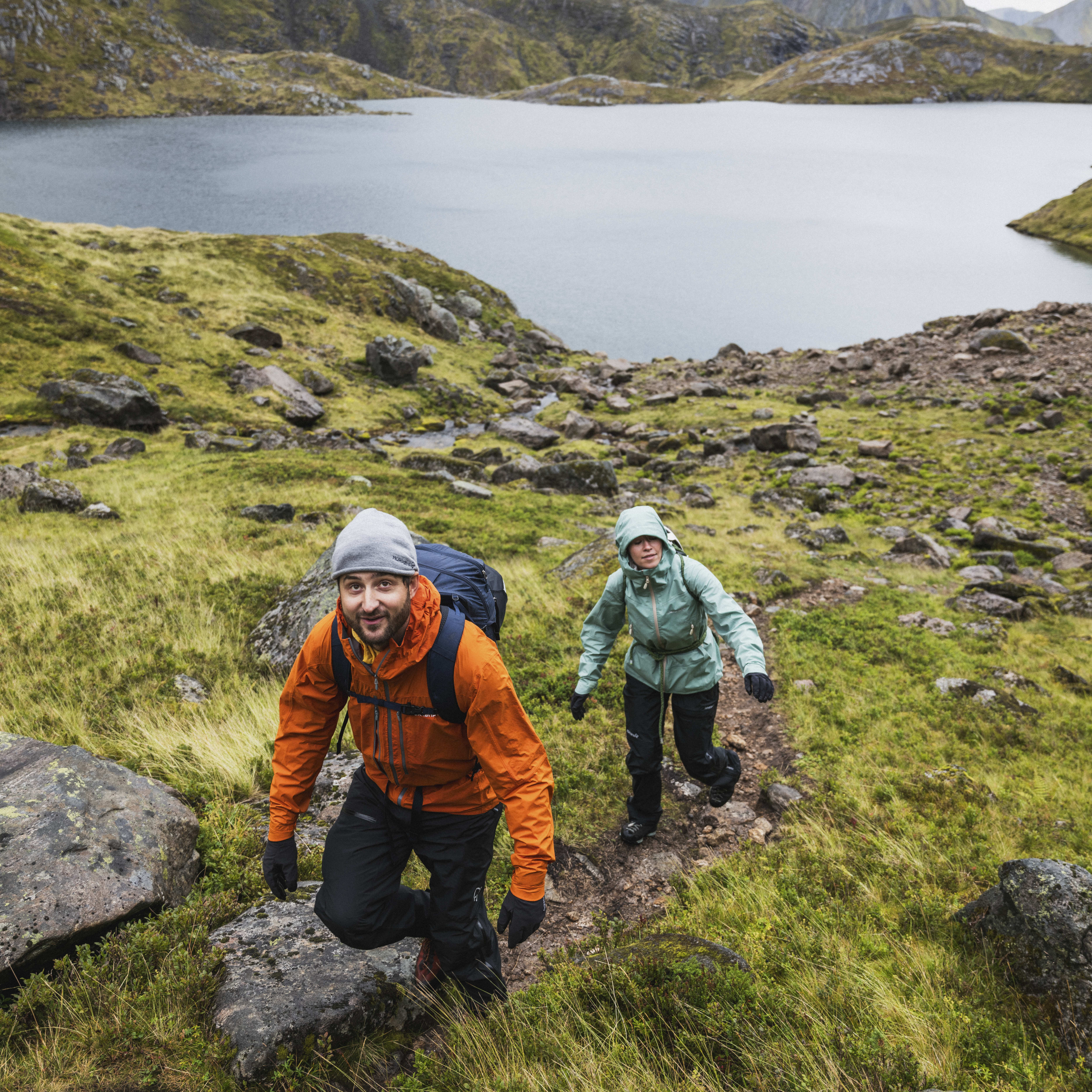 Two hikers wearing Norrøna gear walk a path through grassy terrain with a lake and mountains in the background.