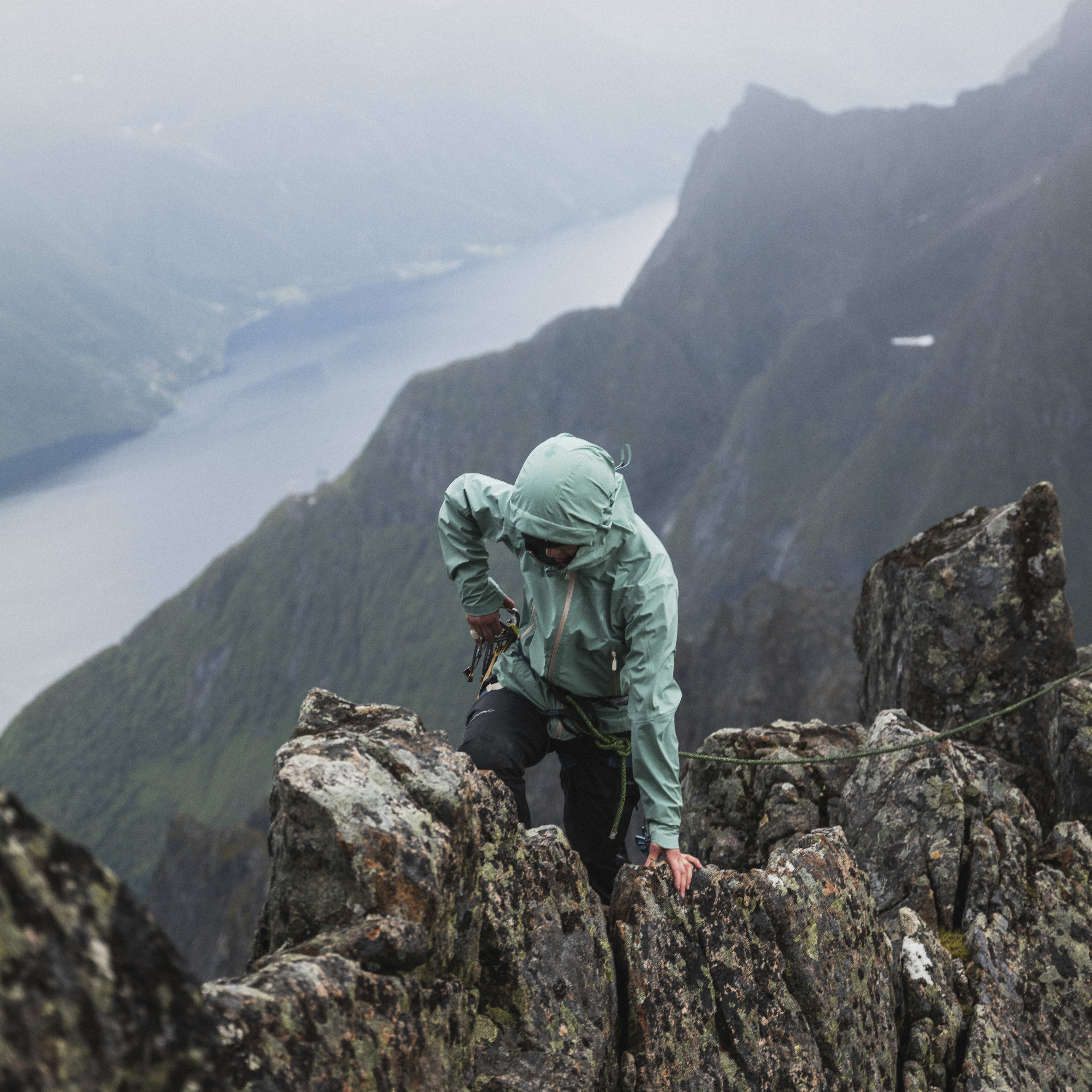 A person wearing Norrøna clothing climbs a rocky mountain ridge. The background shows a fjord and mountains. 

