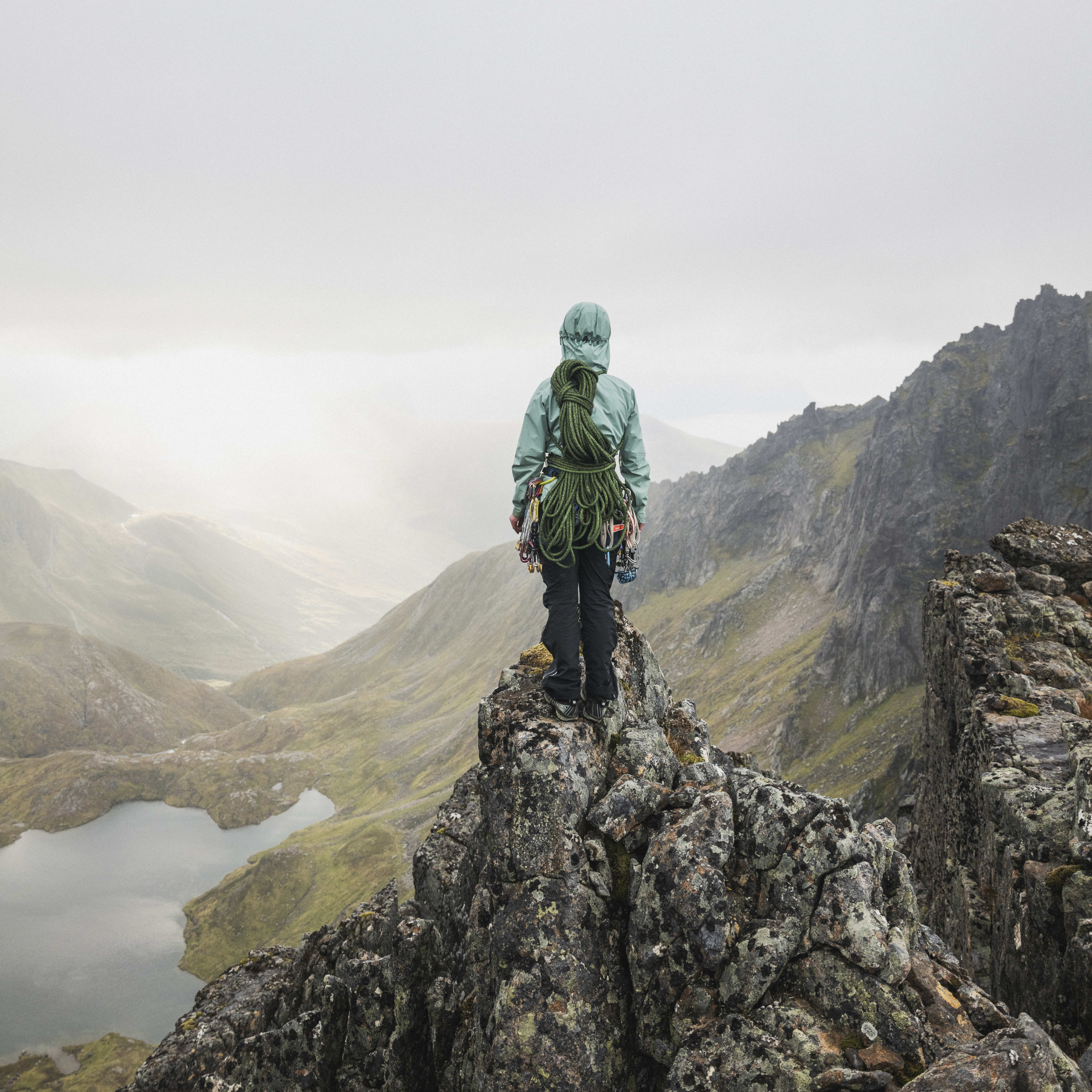 A person wearing a Norrøna jacket and climbing gear stands on a rocky mountain peak overlooking a valley with a lake.