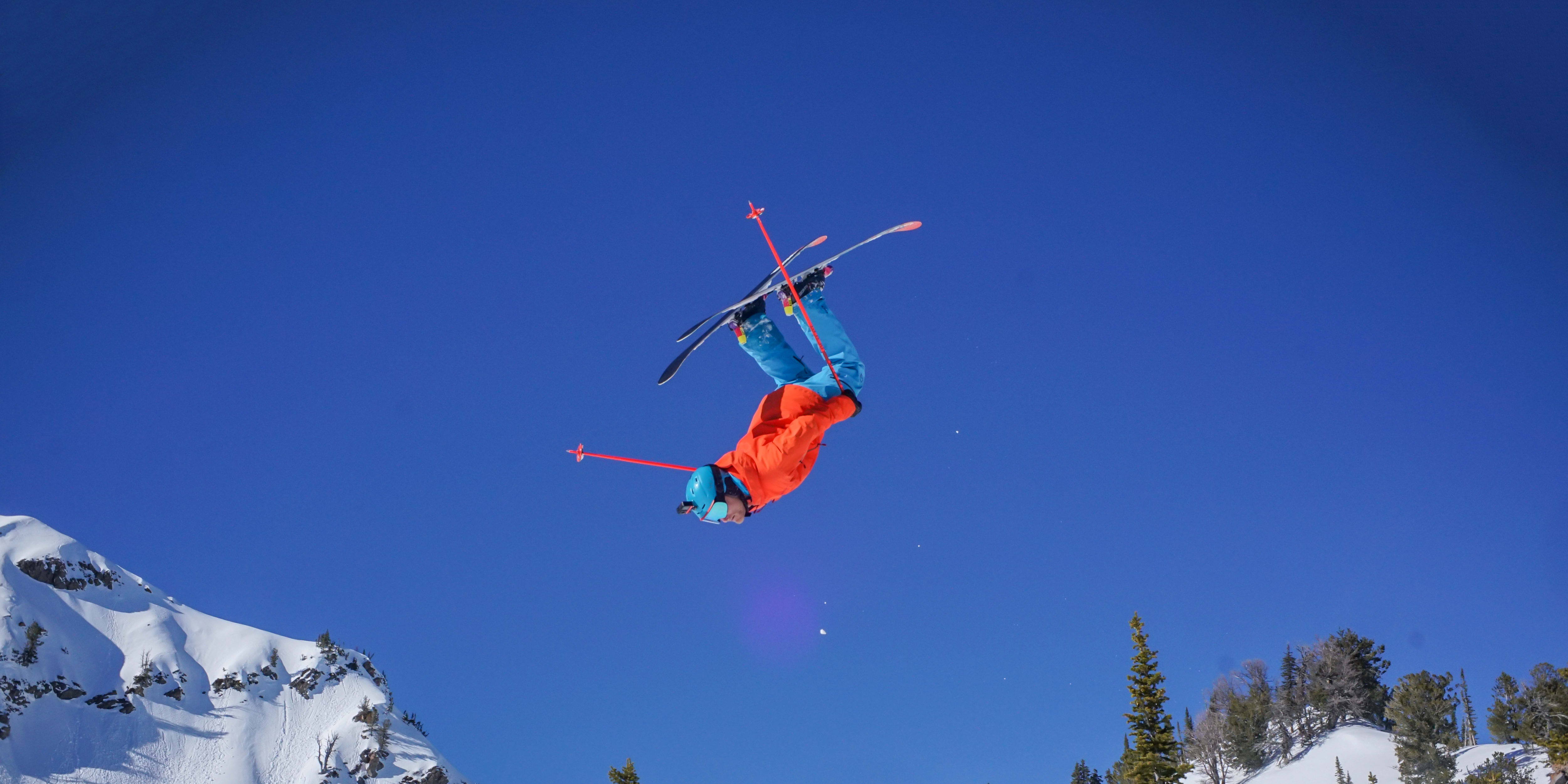 A skier wearing a Norrøna jacket and pants does a backflip against a clear blue sky, with a snowy mountain backdrop.
