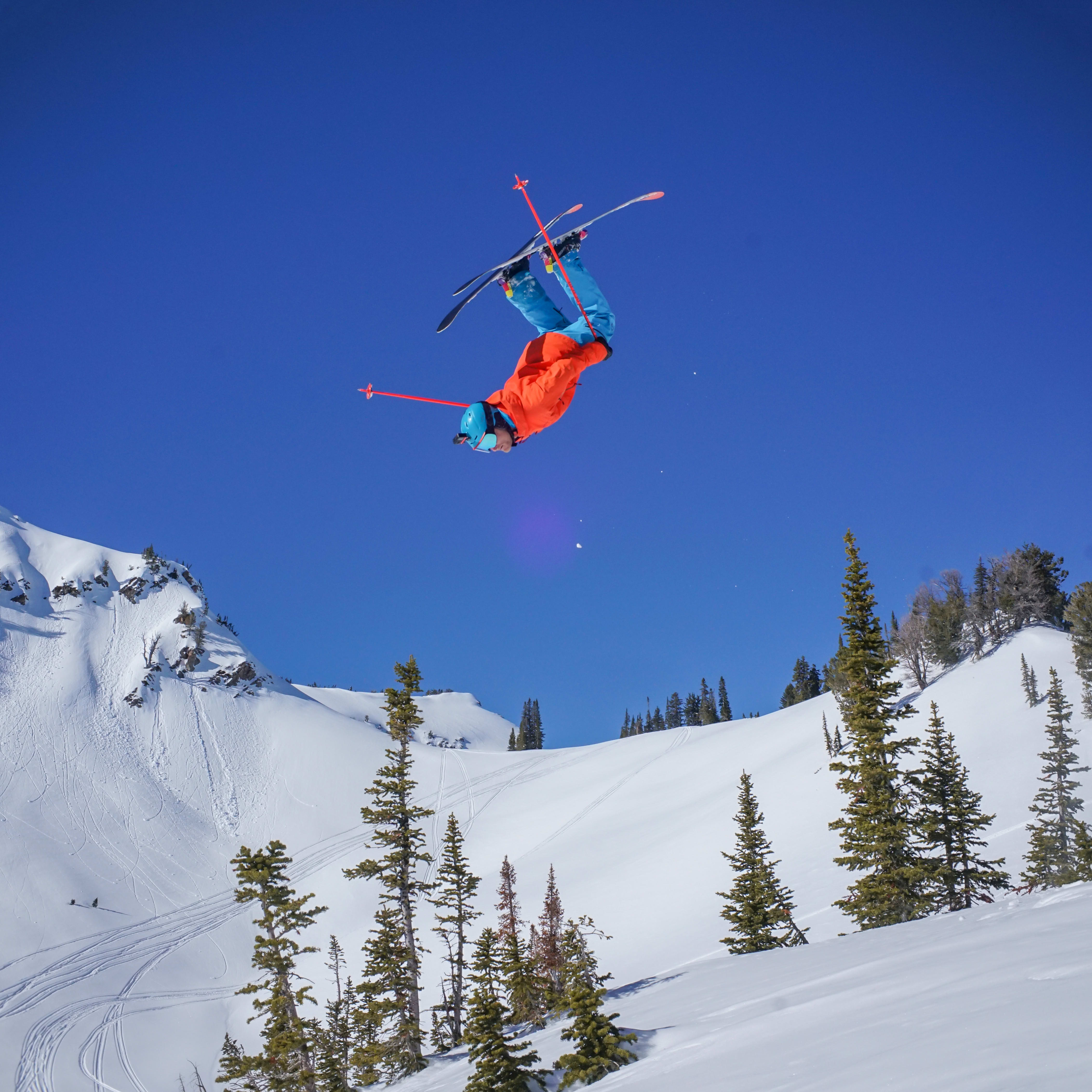 A skier wearing a Norrøna jacket and pants does a backflip against a clear blue sky, with a snowy mountain backdrop.

