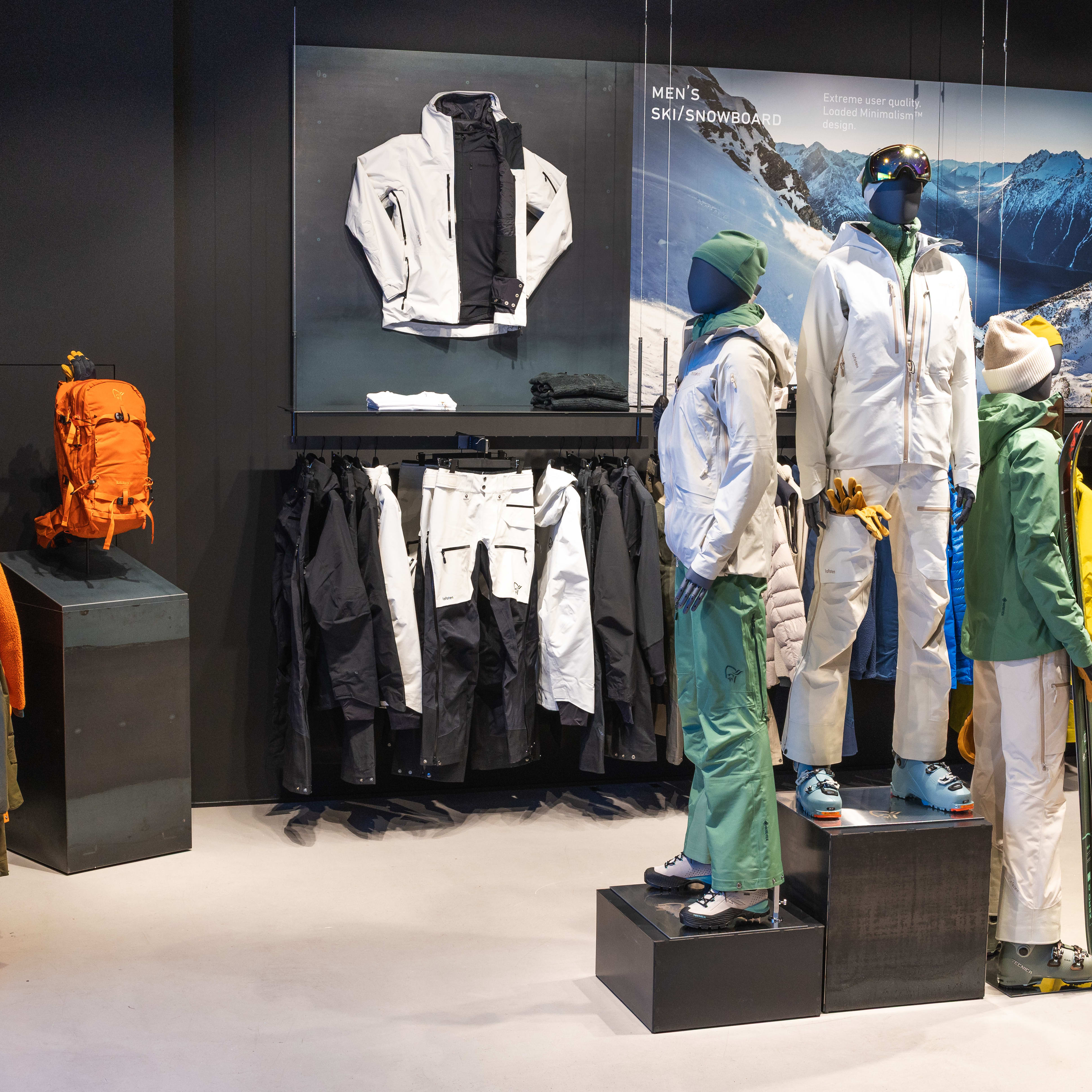 Store display of Norrøna ski/snowboard clothing. Mannequins wear jackets and pants; rack displays more apparel, and an orange Norrøna backpack is featured.