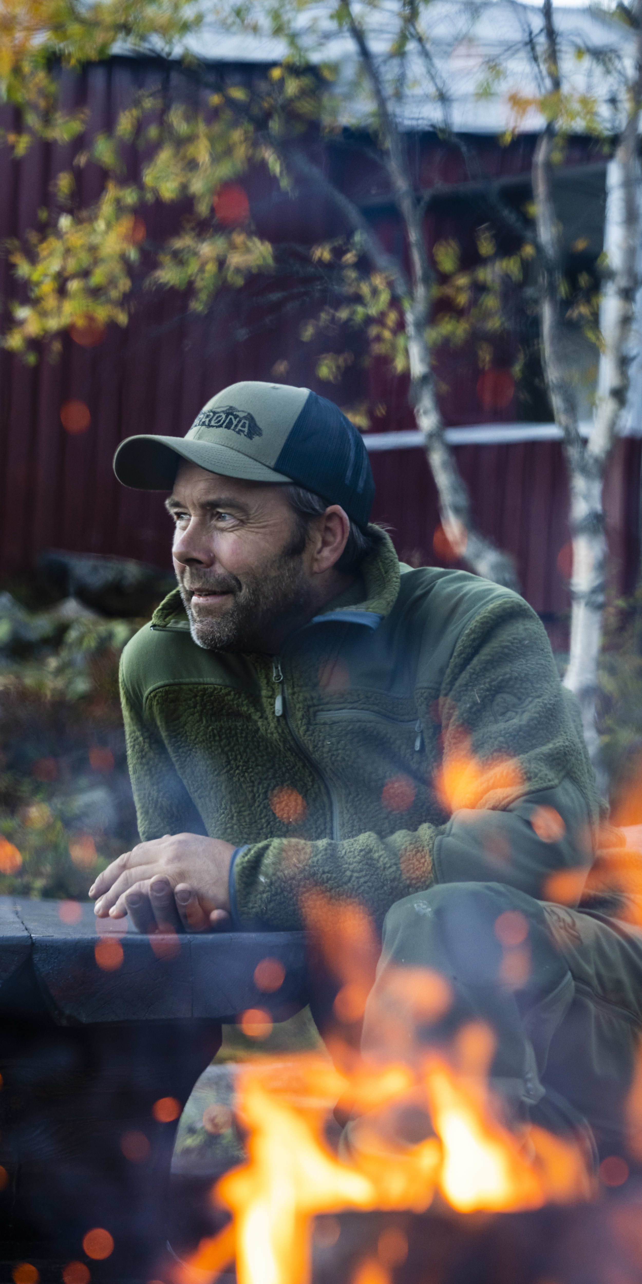 A man wearing a Norrøna jacket sits by a campfire, looking to the side. The fire is in the foreground, with the man blurred out of focus.  

