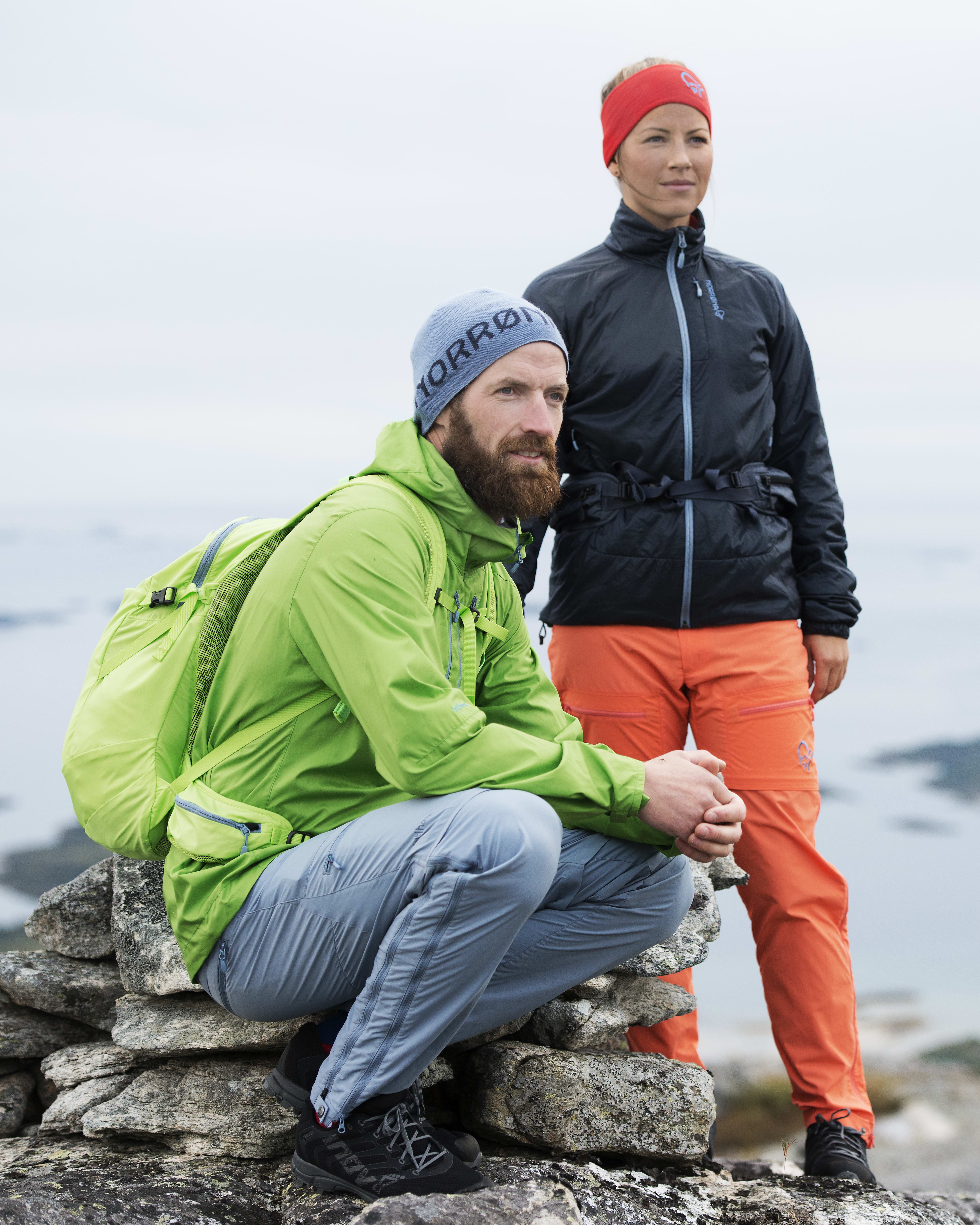 Two people wearing Norrøna clothing sit on a rocky mountaintop overlooking a body of water.  
